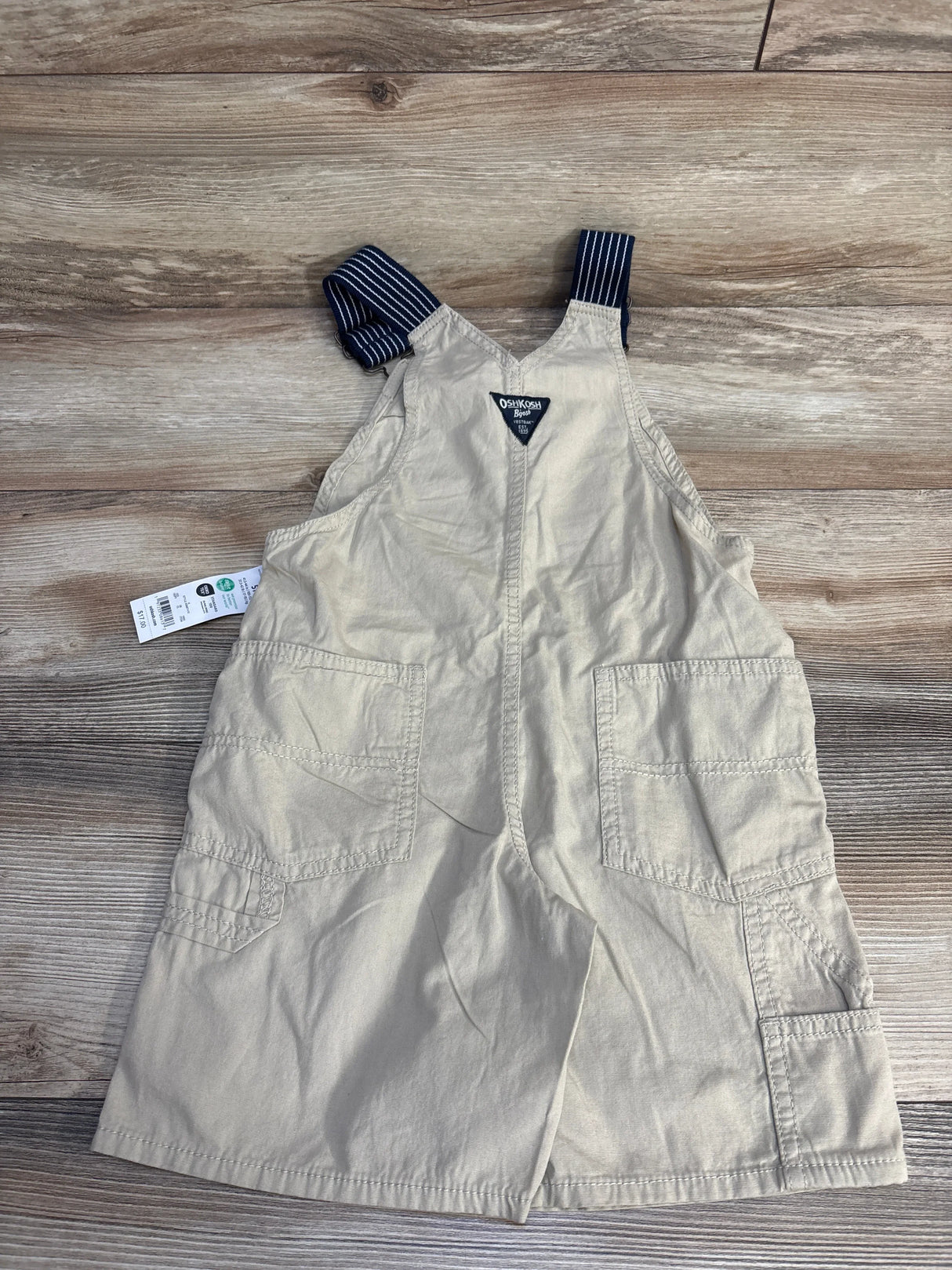 Beige OshKosh B'gosh toddler overalls with navy striped straps, laid on wood floor