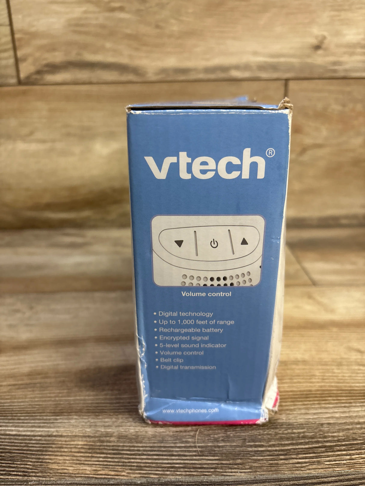 Front view of VTech DM111 baby monitor box showing VTech logo, volume control illustration and feature list on wooden table