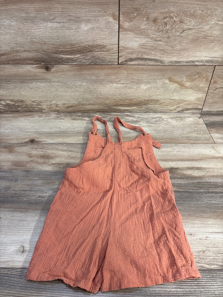 Top-down view of coral gauze shortalls showing crinkled gauze texture and knotted shoulder straps on light wood background