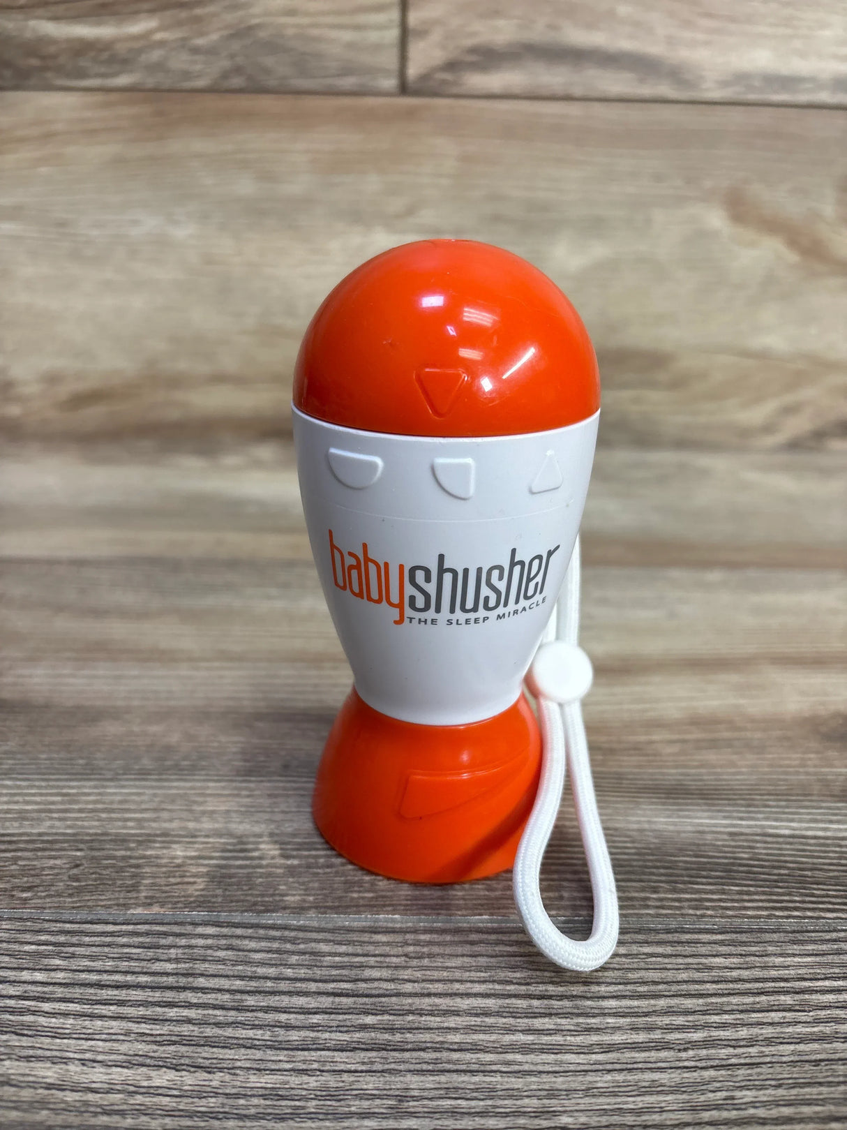Front view of Baby Shusher sleep soother sound machine showcasing orange and white casing and attached white wrist strap on wood surface