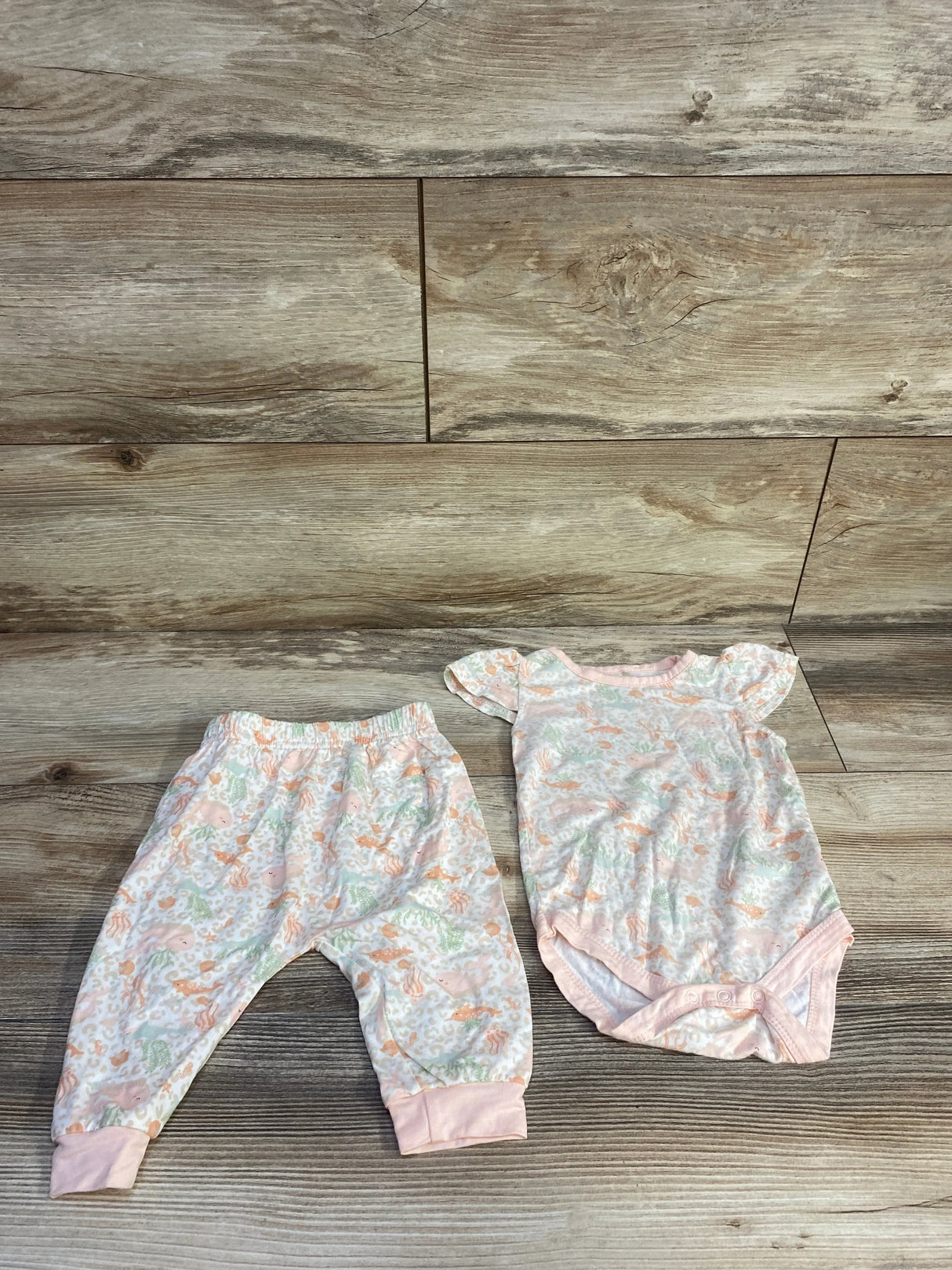 Flat lay of Milkberry 2-piece sea animal print bodysuit and pants in pale pink/white showing flutter sleeves, snap crotch, elastic waist on wood background