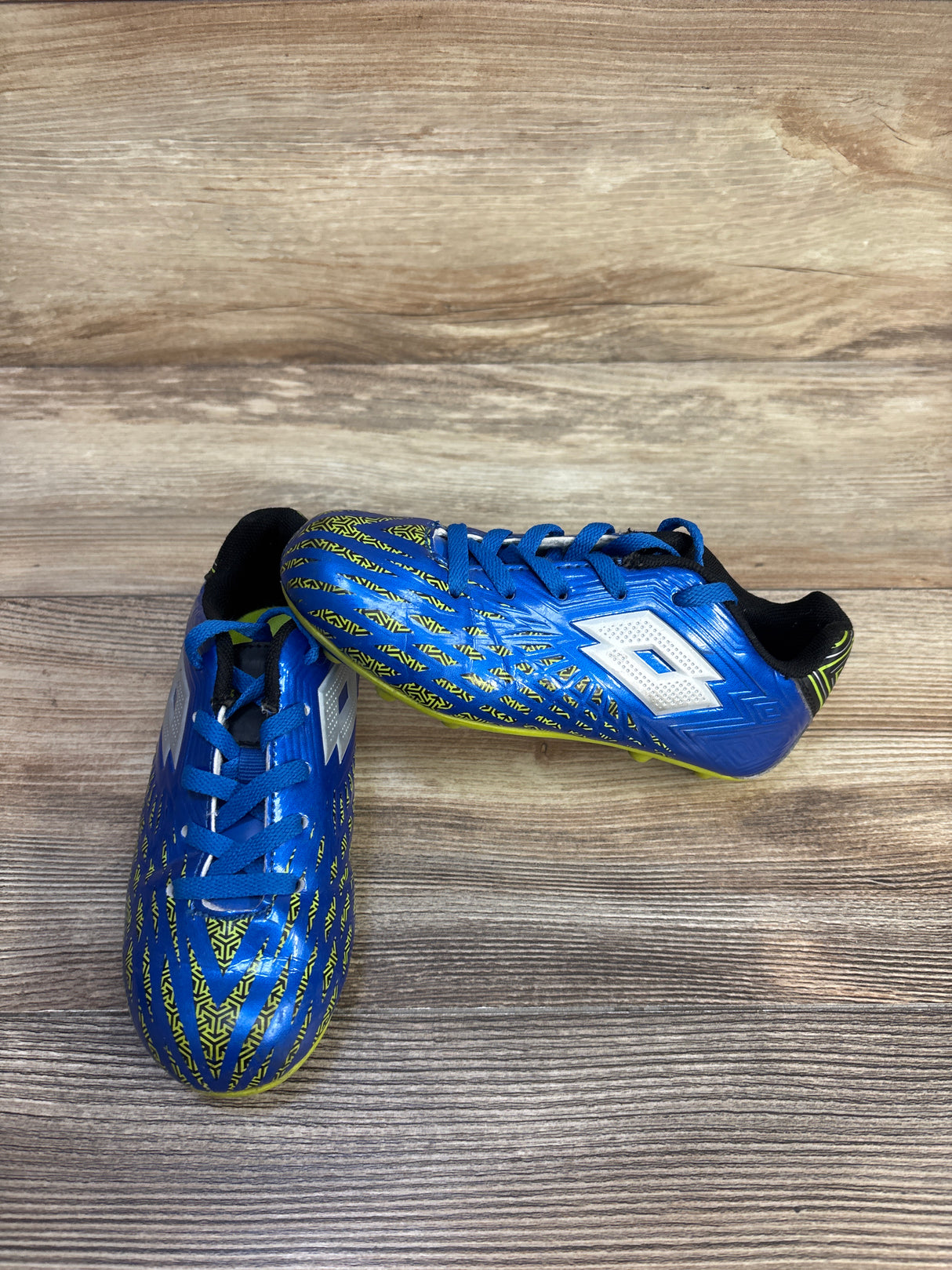Three-quarter view of junior blue Lotto Forza II cleats showing patterned upper, Lotto logo, and studs on wood background