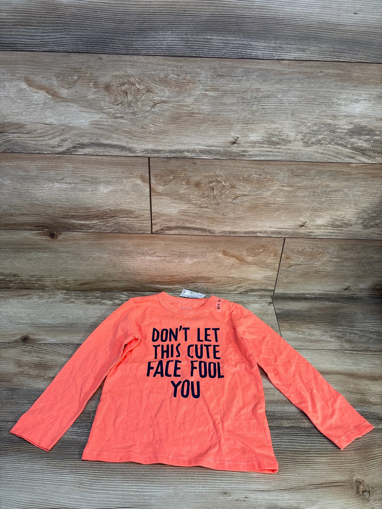 Long sleeve orange kids shirt with 'Don't let this cute face fool you' text, laid on wood floor