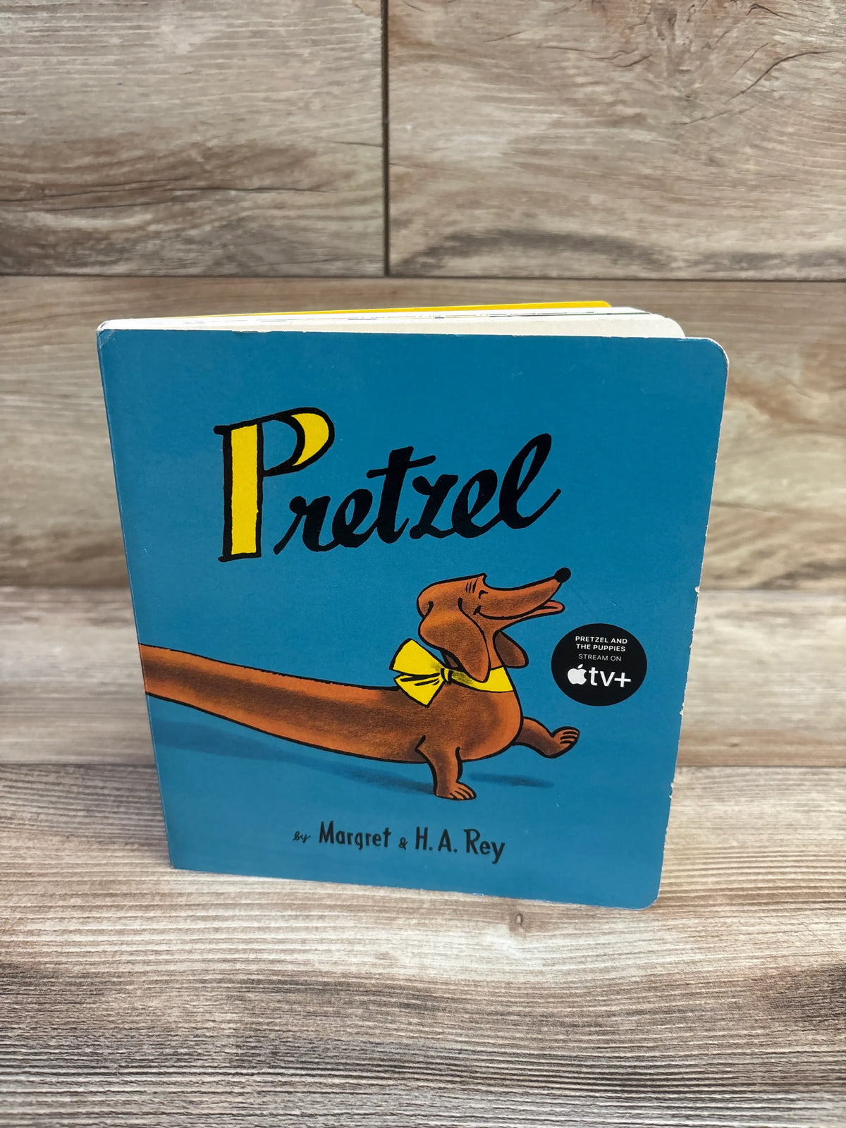 Front view of Pretzel board book with blue cover, illustrated dog in yellow bow tie, on a wood-grain background