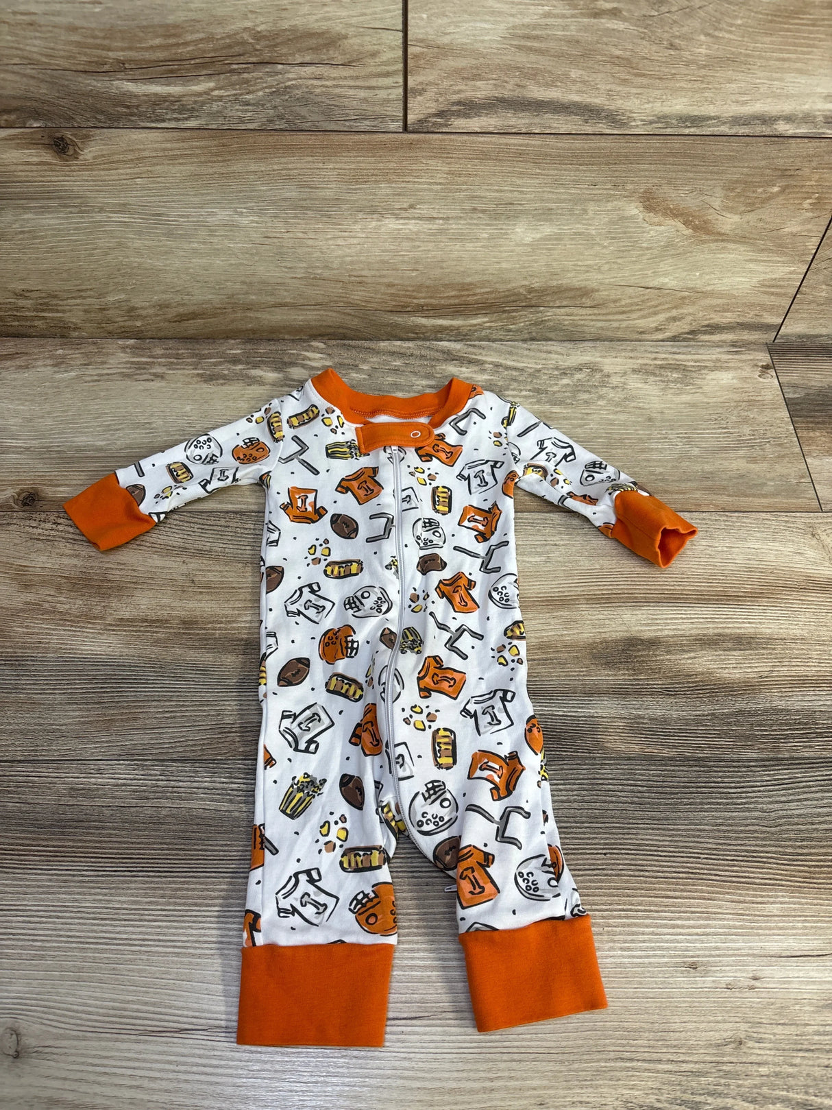 Top-down view of Magnolia Baby white footless sleeper with orange cuffs and football-themed print, front zipper on wood background
