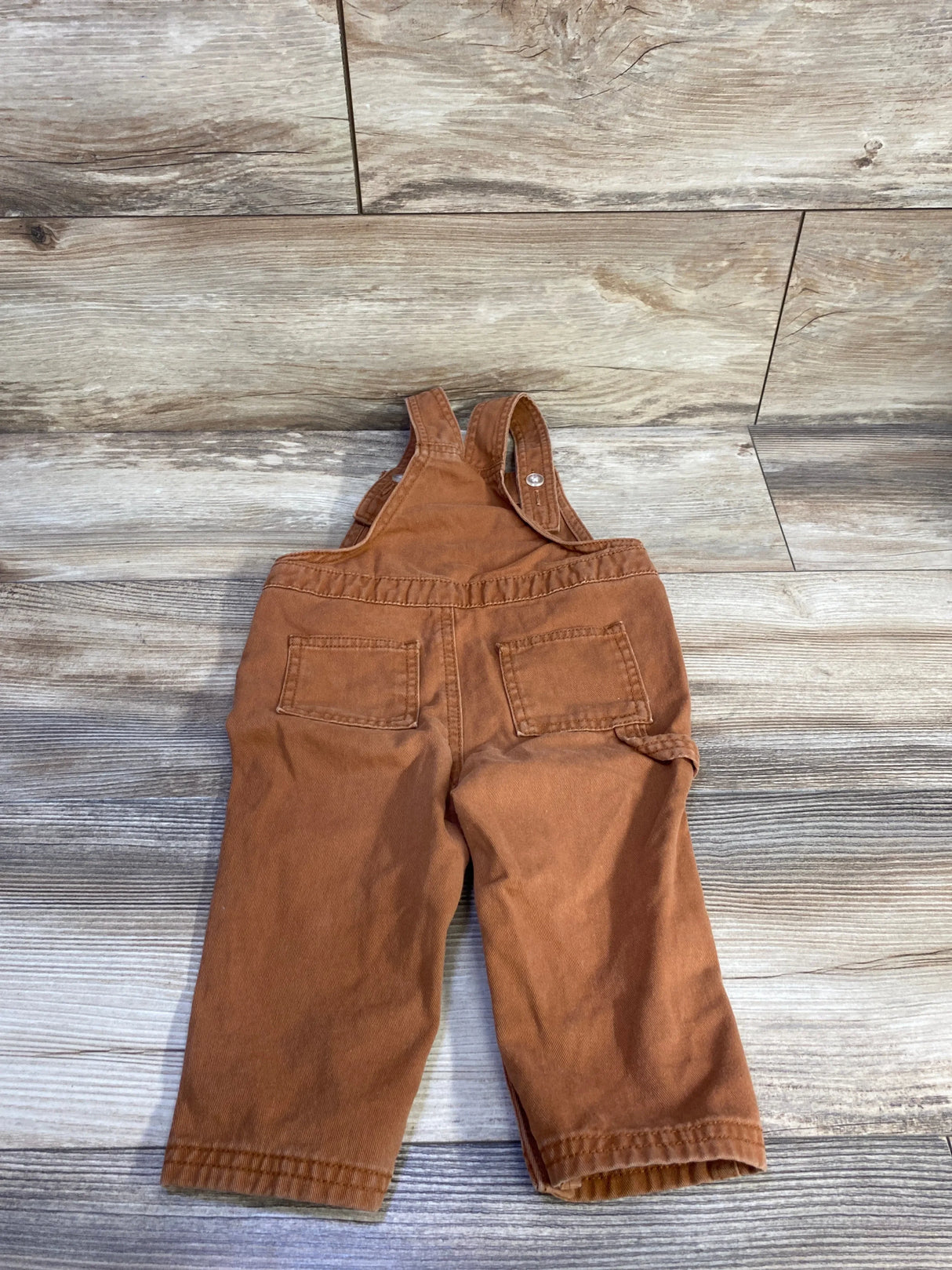 Back of brown twill Old Navy toddler overalls, 12-18m, showing back patch pockets and adjustable straps on wood floor
