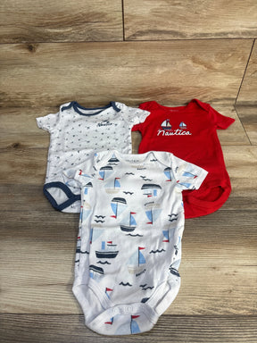 Top down view of three Nautica baby bodysuits on wood floor: white/navy, red with sailboat logo, white sailboat print