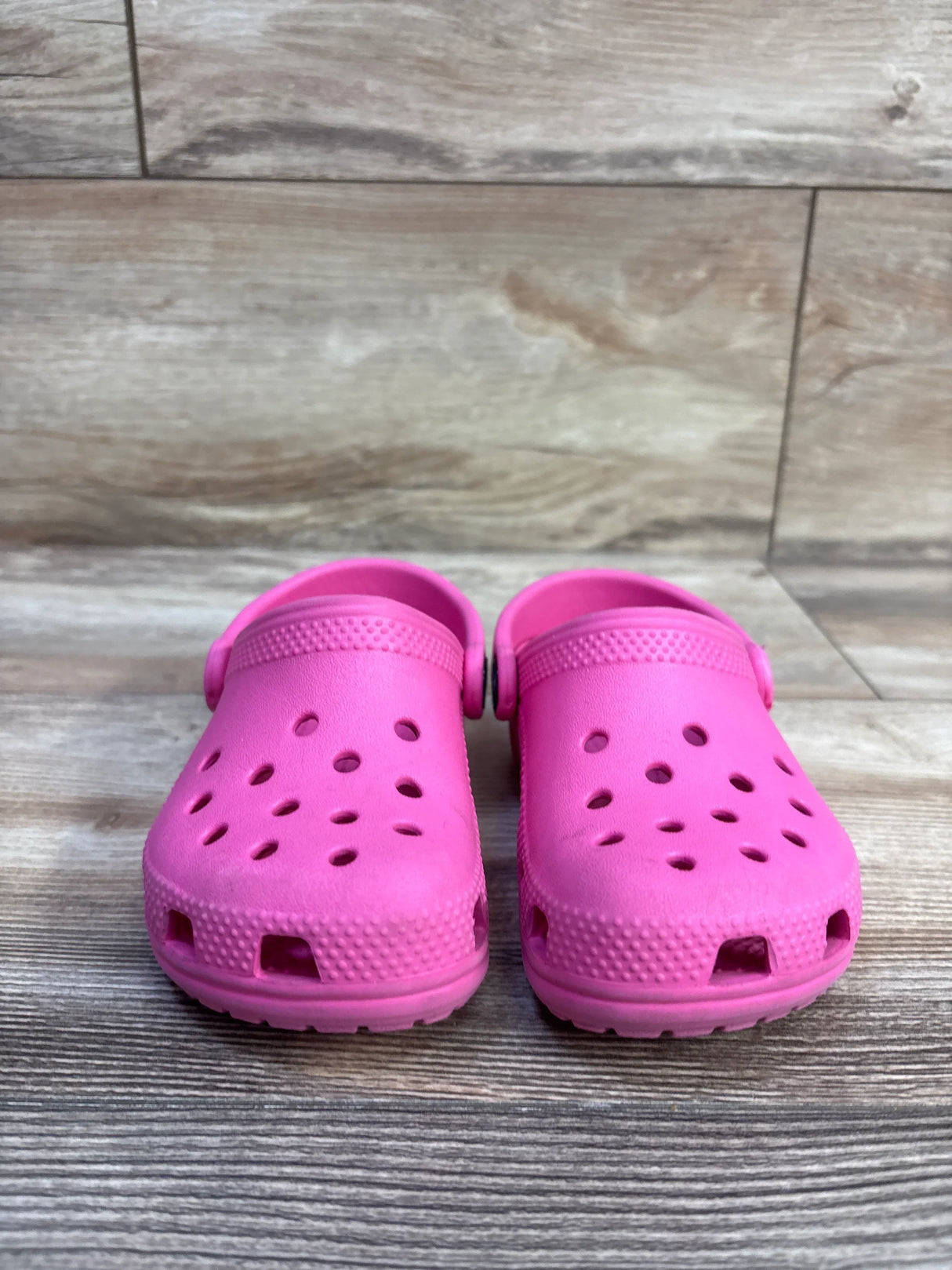 Front view of pink Crocs Classic clogs showcasing ventilation holes, textured toe bumper, and heel straps with wood-look floor background