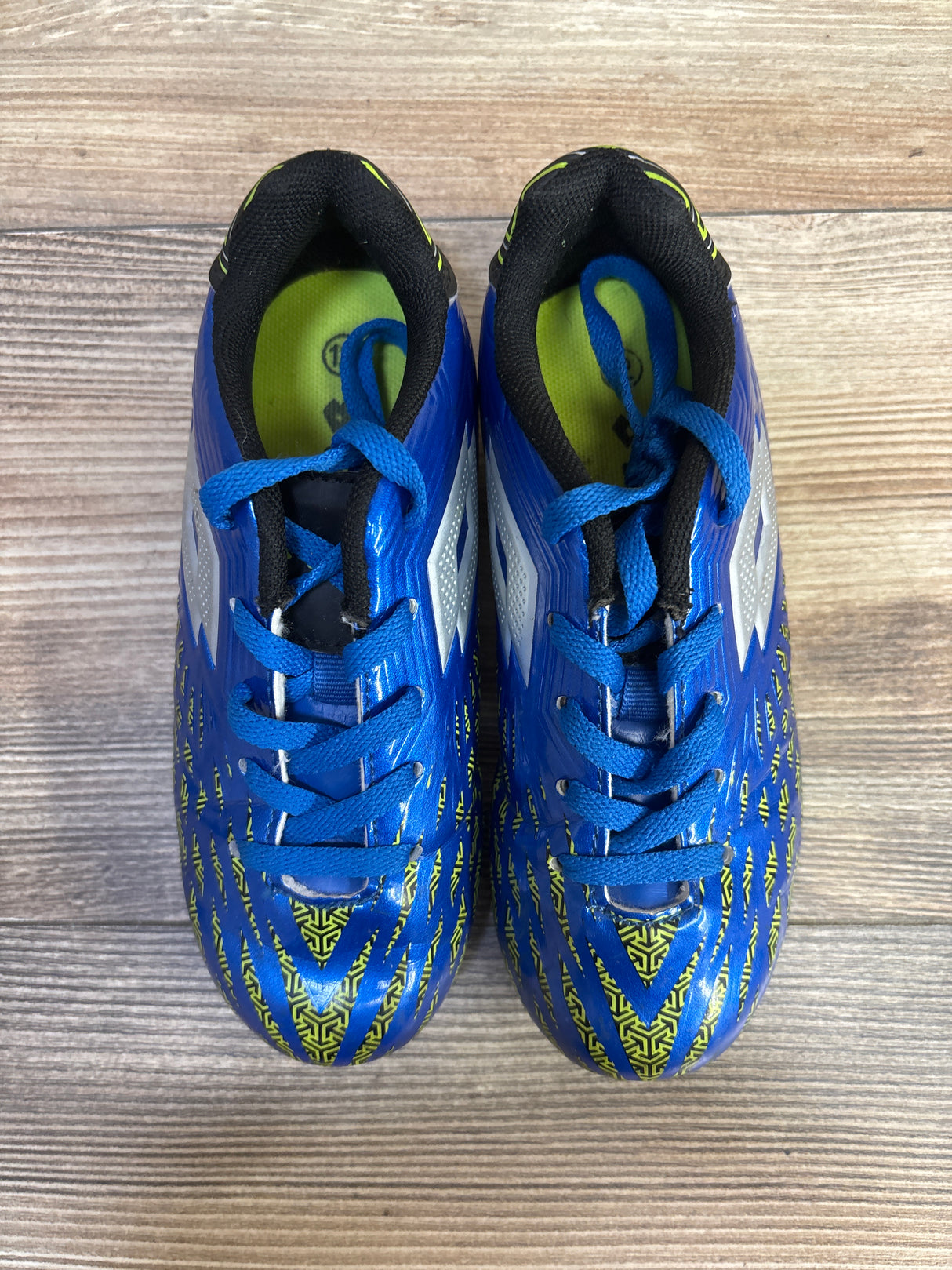 Top-down view of Lotto Forza II Jr soccer cleats in blue showing patterned yellow-green toe, white side logos, and lace-up closure on wood floor