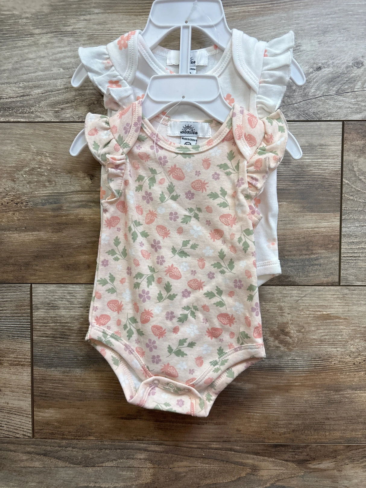Front view of two Wonderluxe flutter sleeve bodysuits, pink strawberry print and white, 0-3M, on hangers on wood floor