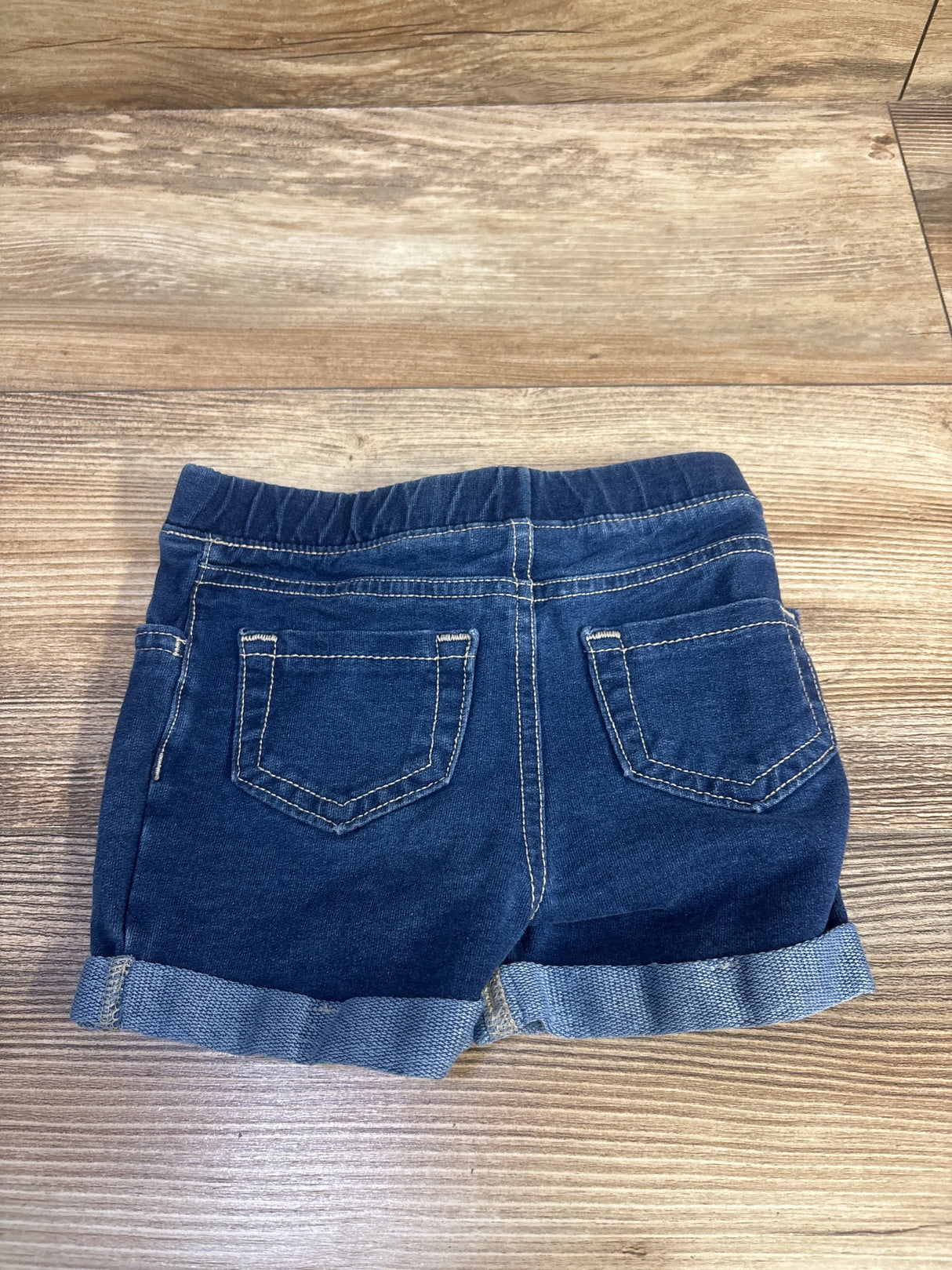 Back view of Jumping Beans pull-on blue denim toddler shorts size 18m showing elastic waistband, cuffed hems, and back pockets on wood surface
