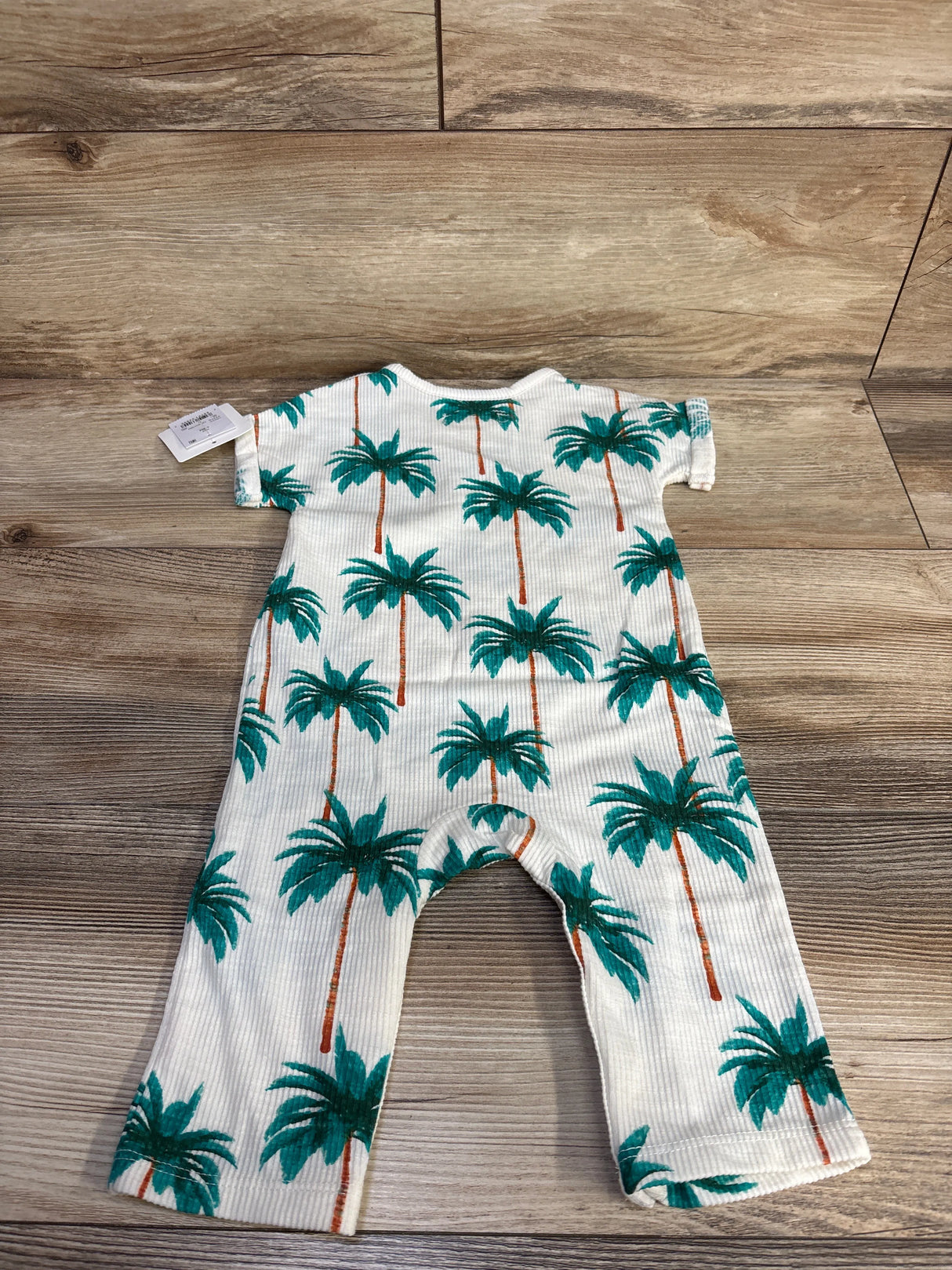 Top-down view of white Grayson Mini ribbed baby jumpsuit size 6-9 months showing teal palm tree print on wood background