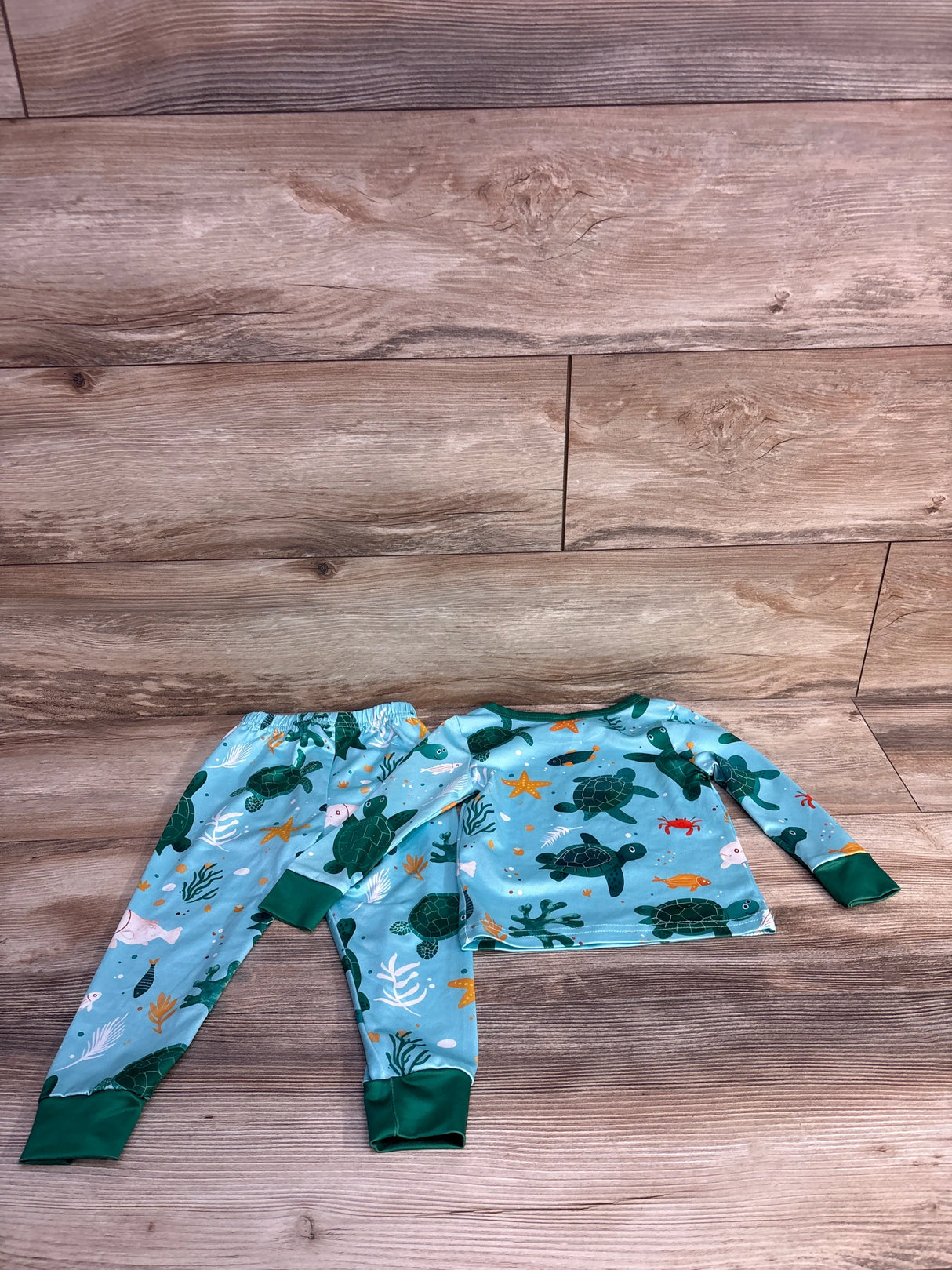 Flat lay of blue 2-piece sea turtle 2T pajamas showing long-sleeve top and elastic pants with green cuffs on wood floor