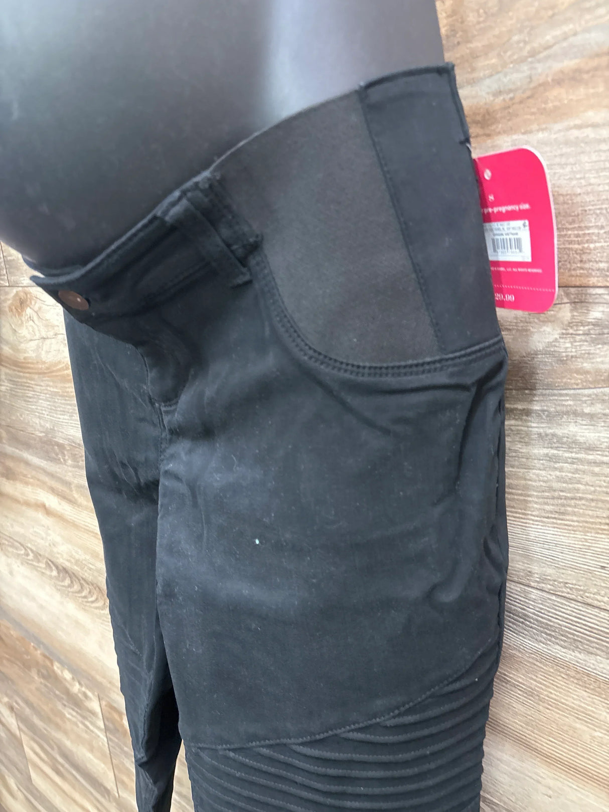 Close-up side view of black maternity utility jeggings showing elastic over-belly panel, front pocket and ribbed knee detail on wood floor background
