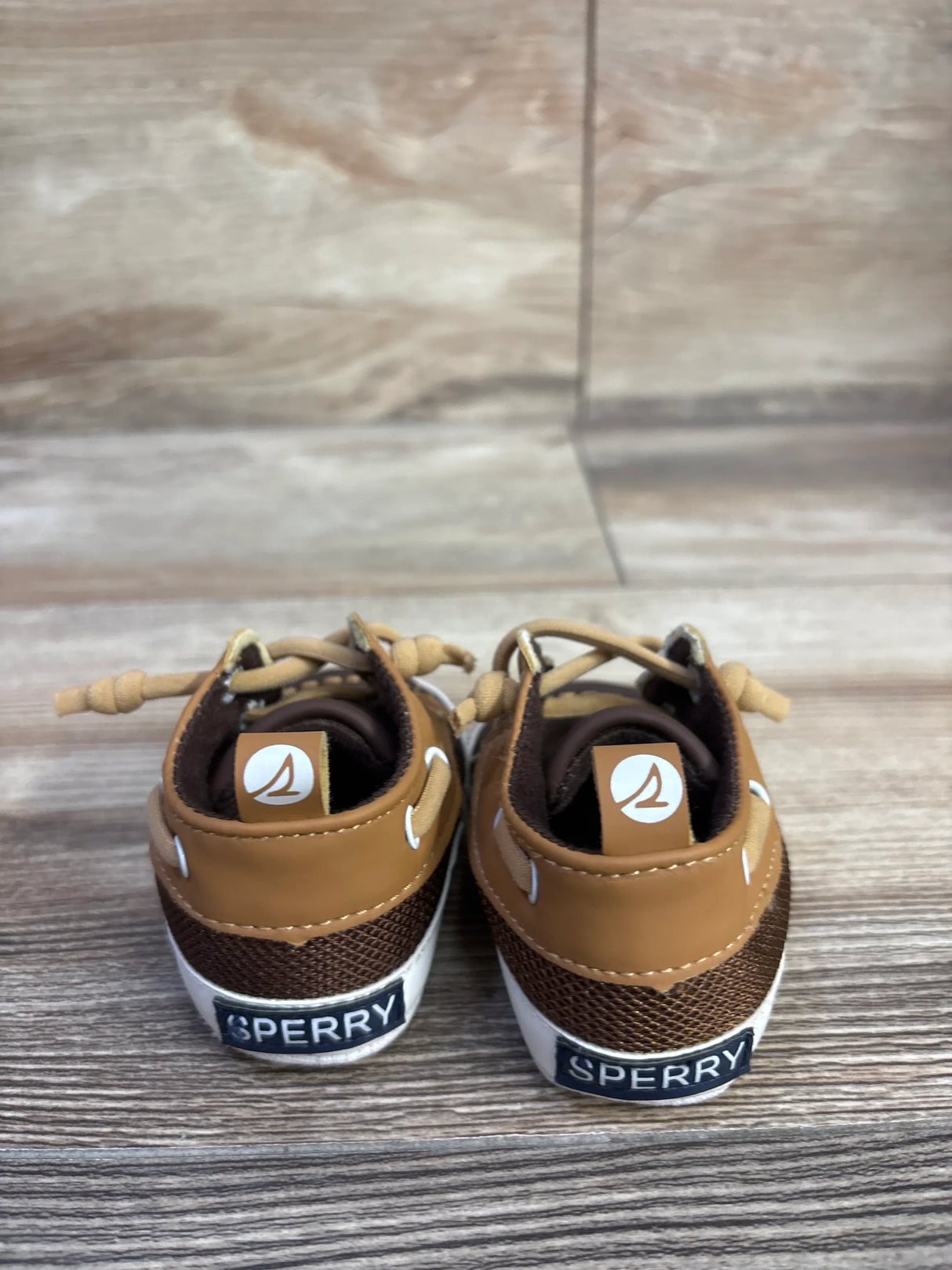 Rear view of Sperry baby boat crib shoes showcasing tan leather and brown mesh uppers with white soles on wood background