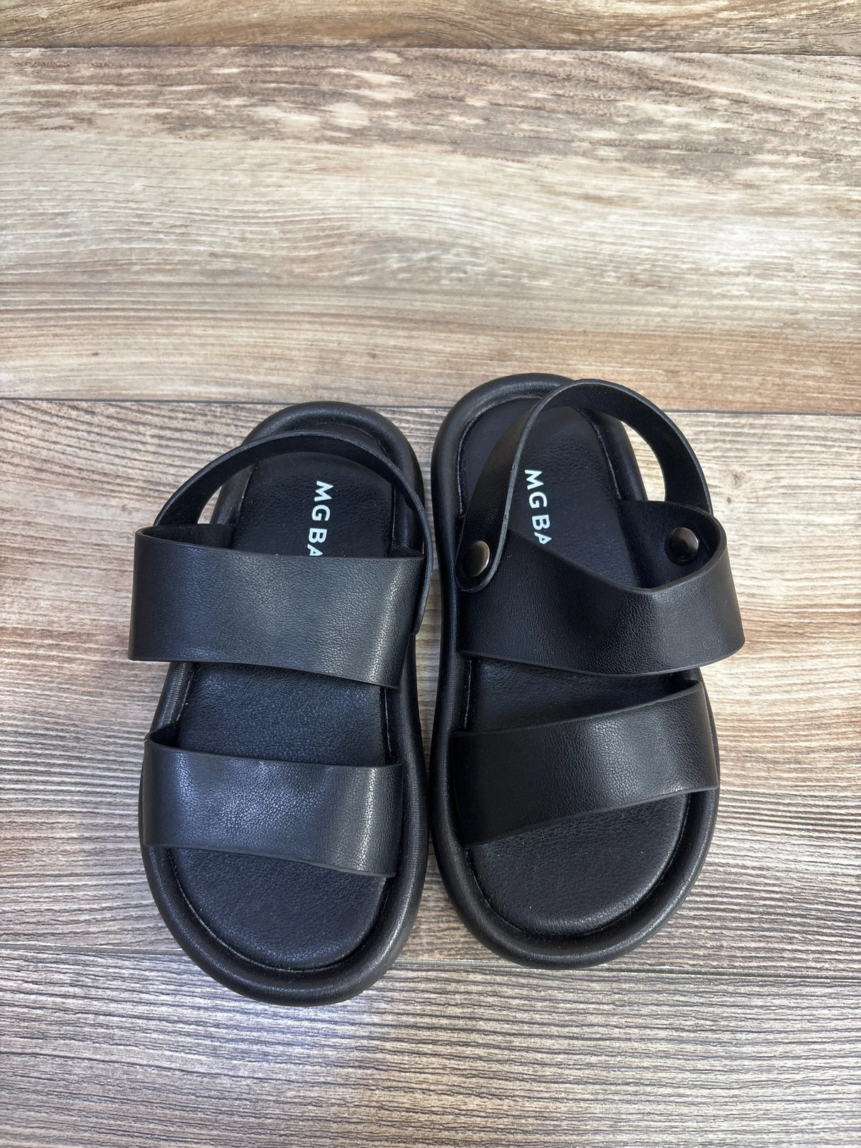 Top-down view of black MGBABY foam toddler sandals showing two wide straps and adjustable snap-back on wood floor
