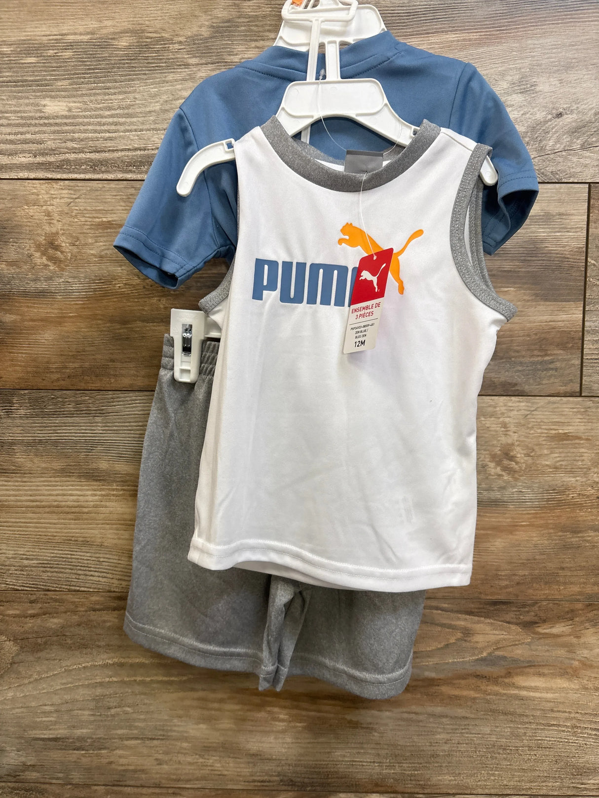 Front view of 3-piece baby Puma set: white tank with blue PUMA text and orange puma logo, blue tee, and gray shorts on wooden background