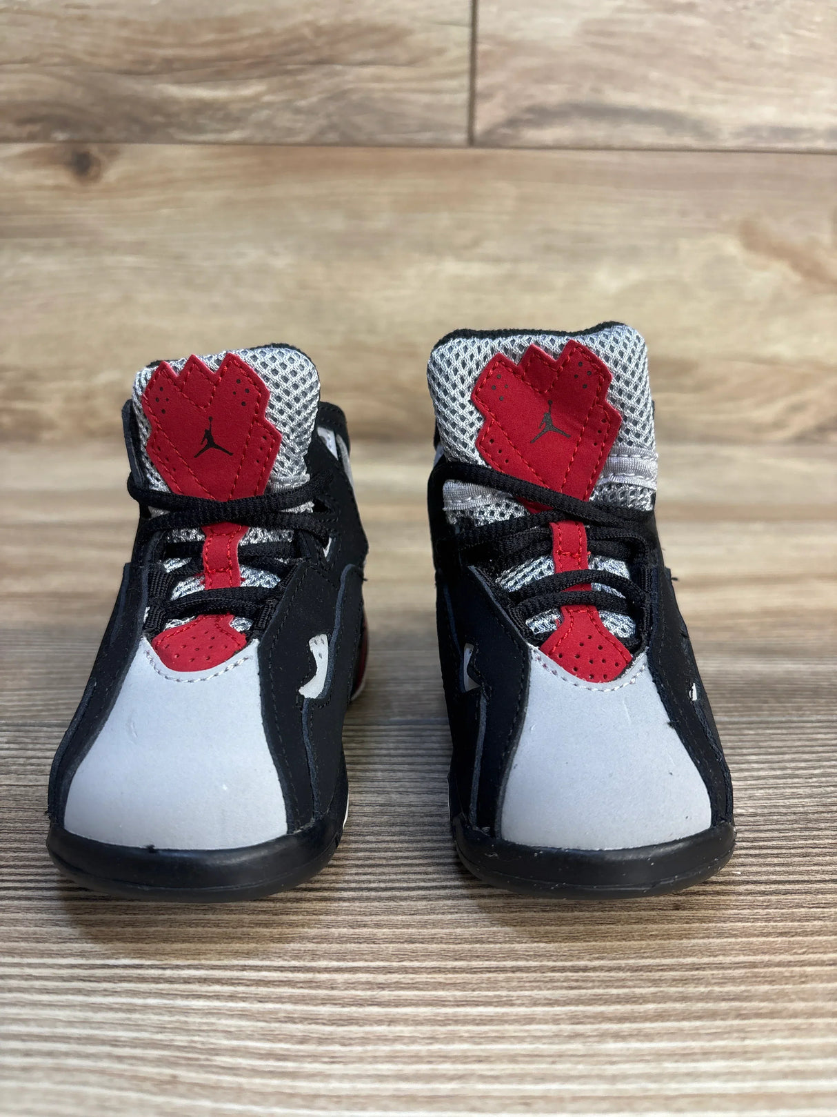 Front view of Air Jordan True Flight TD toddler sneakers in black, red and grey showing mesh tongues, Jumpman logo, and padded ankle on wood floor