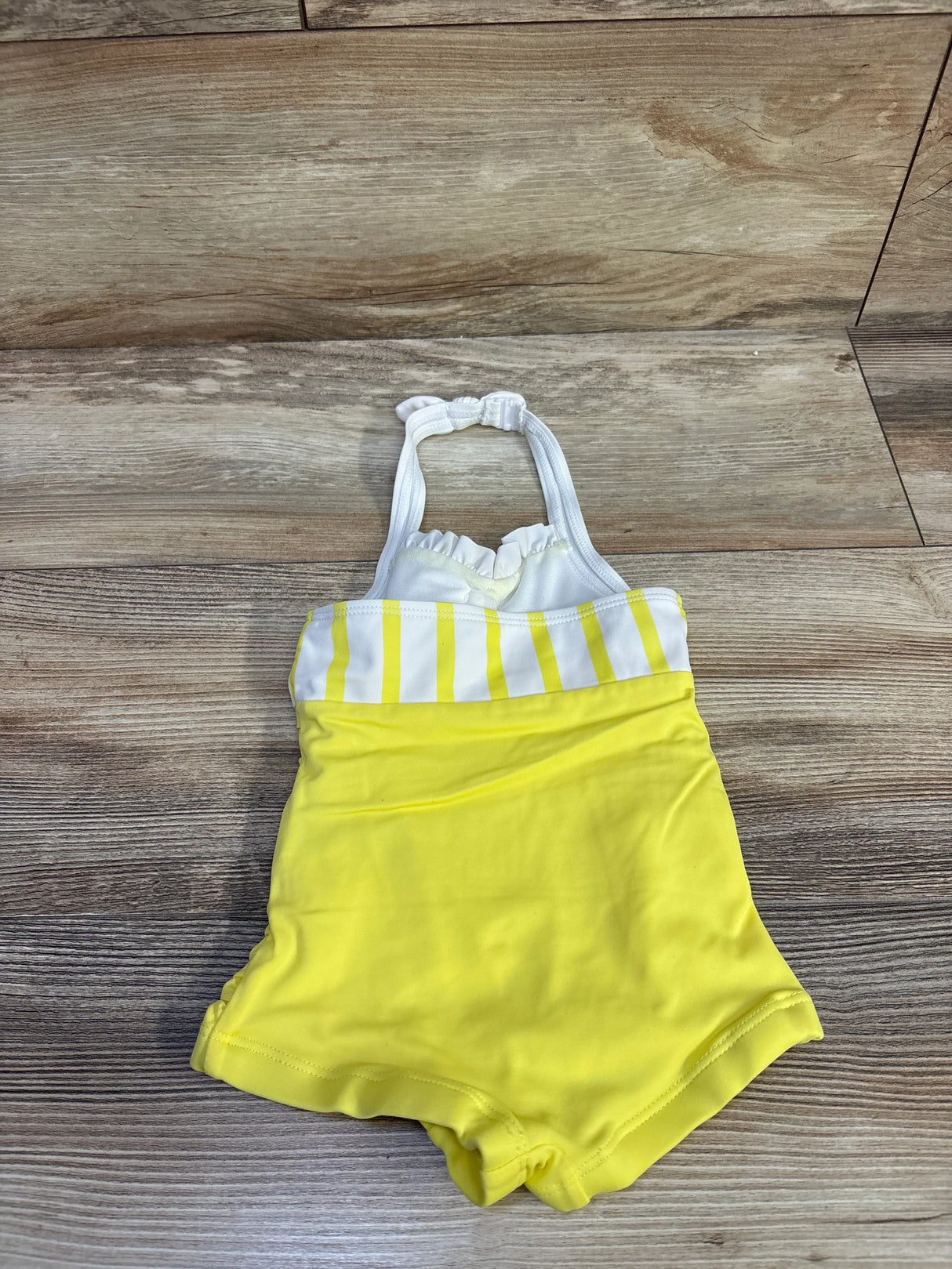 Flat lay of yellow Janie & Jack toddler halter one-piece swimsuit showcasing white striped bust and ruffled halter straps on wood background