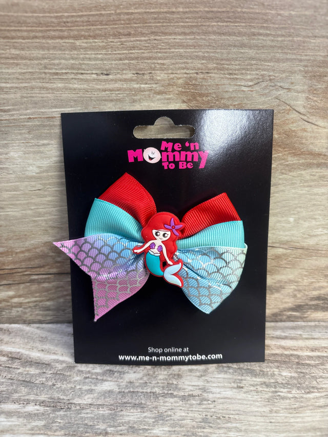 Front of mermaid hair bow showcasing red and teal ribbons, scale-pattern tails, and mermaid center on black display card