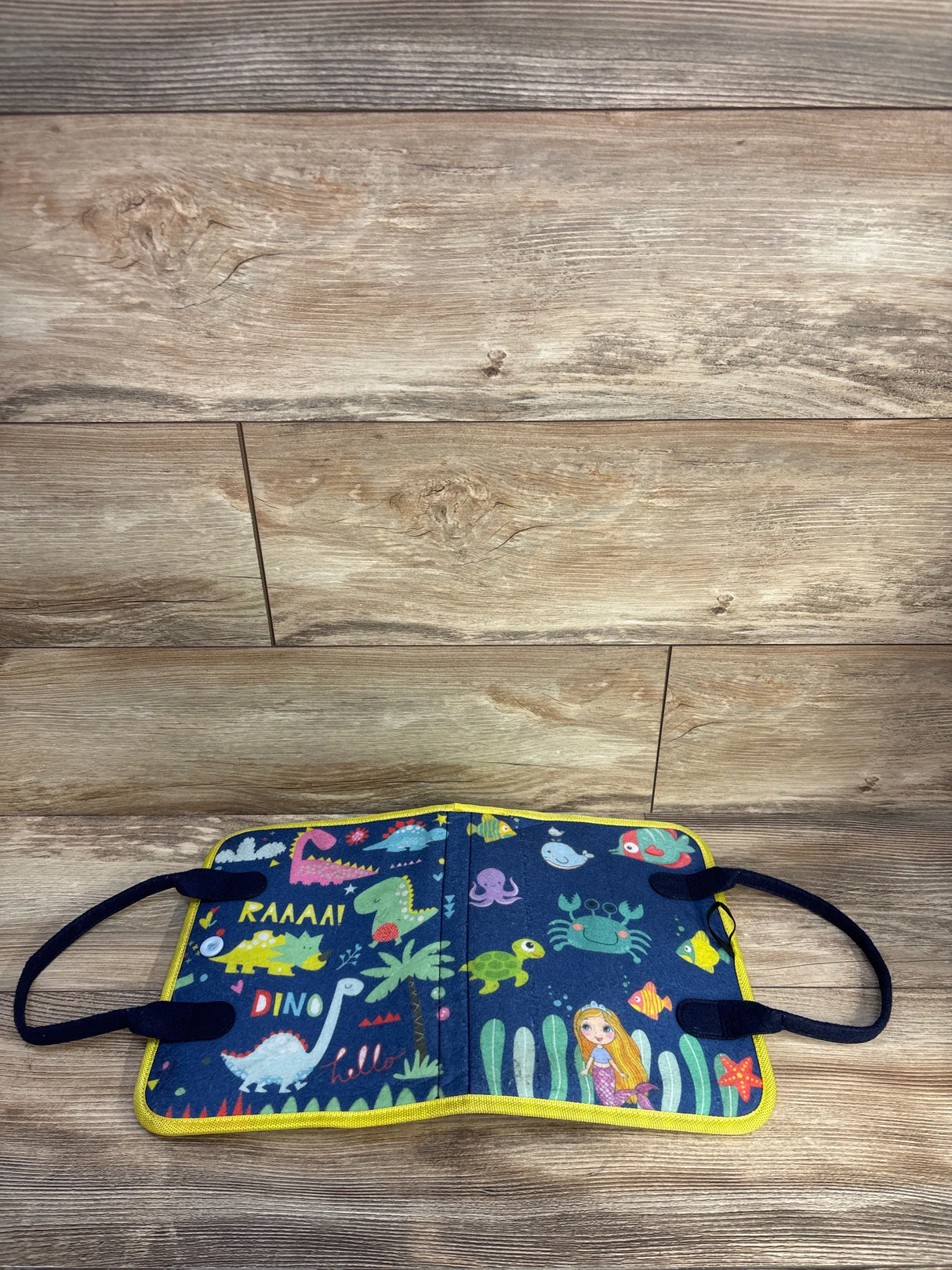 Top-down view of Hooku busy felt book opened showing colorful dinos and sea creatures, elastic straps and yellow trim on wood