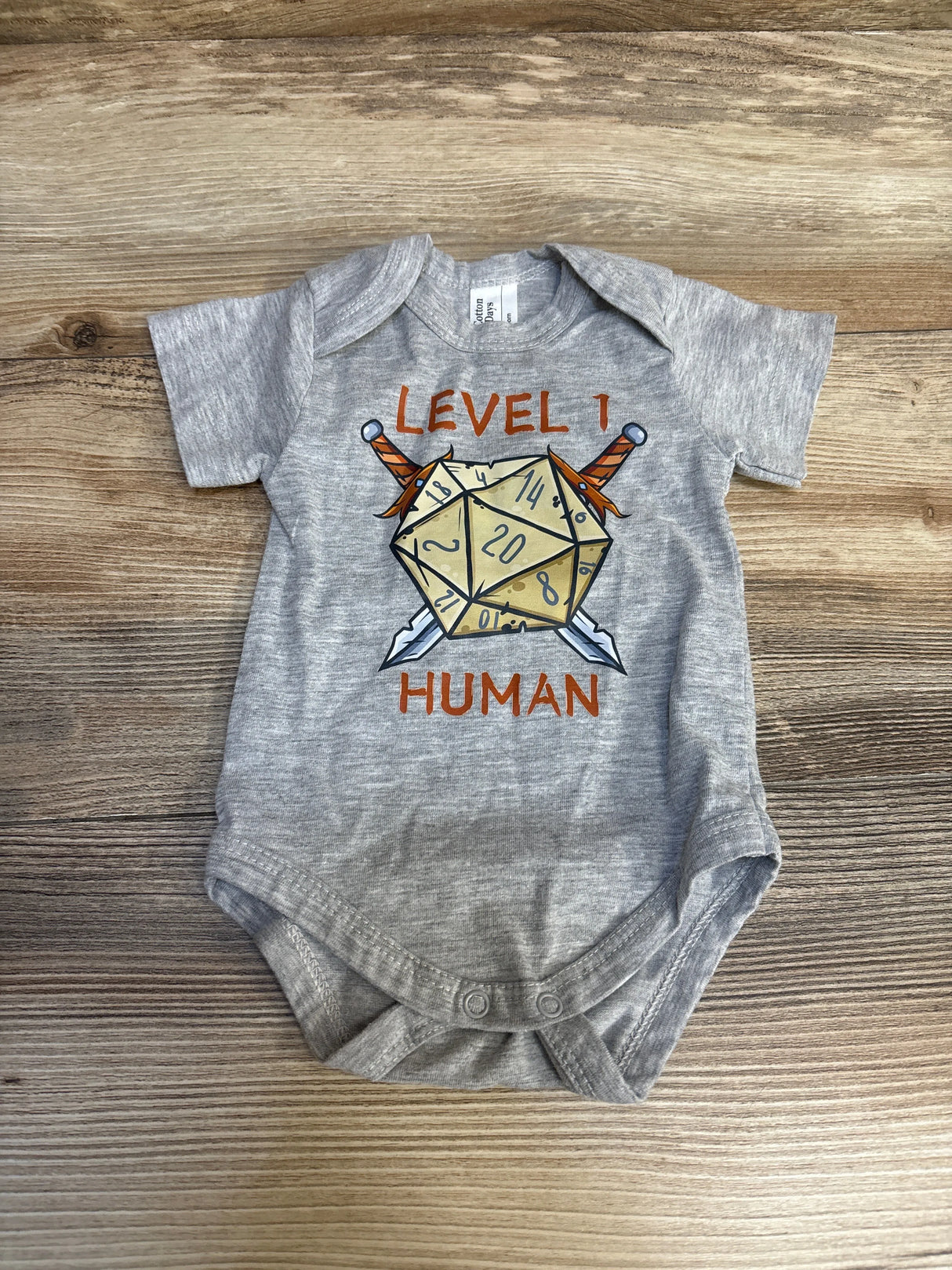 Gray baby onesie with D20 dice, swords, and 'Level 1 Human' graphic, laid on wood background