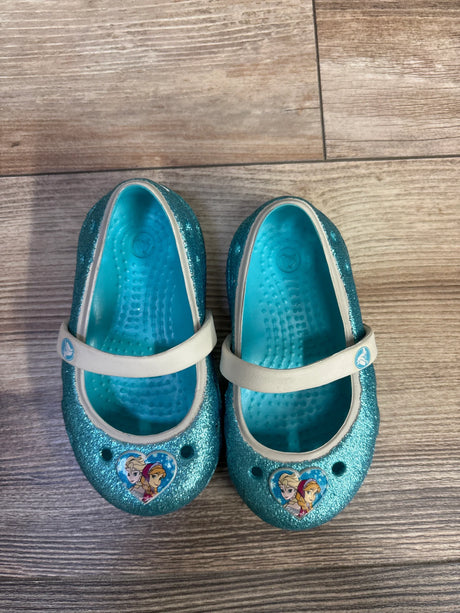 Top view of toddler Crocs Mary Jane flats, blue glitter finish, Elsa and Anna heart charms on toes, white straps on wood floor