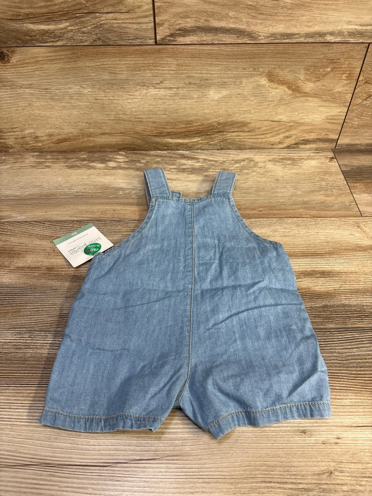 Top-down view of light blue chambray baby shortall sized 6 months showing shoulder straps and center seam on wood background