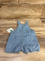 Top-down view of light blue chambray baby shortall sized 6 months showing shoulder straps and center seam on wood background
