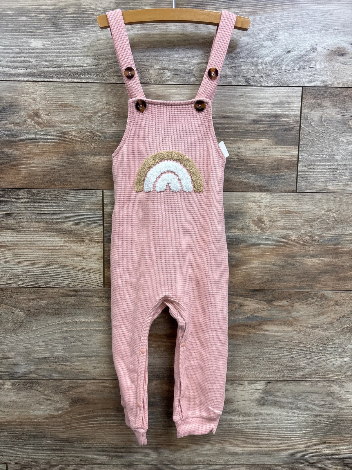 Front view of pink waffle-knit toddler overalls with rainbow applique and button straps on wooden hanger