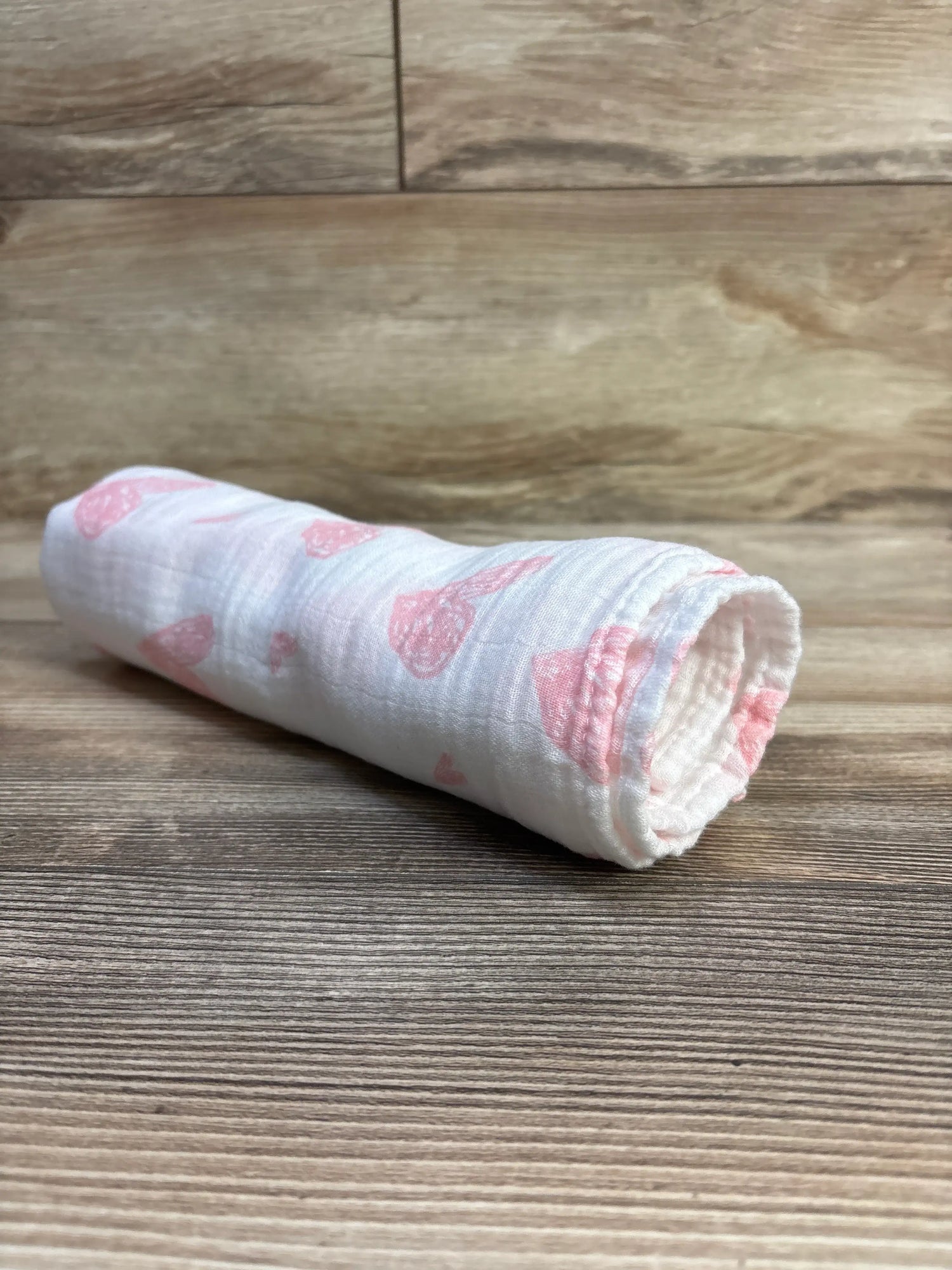 Angled view of a rolled white GLLQUEN muslin bamboo swaddle blanket showcasing pink printed motifs on a wood surface