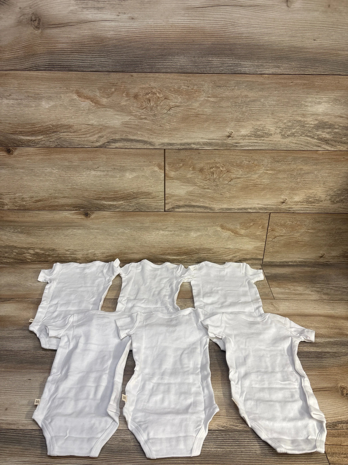 Flat lay of six white Huggies short-sleeve baby bodysuits (3M) showing front snap closures and crew necks on a wood background