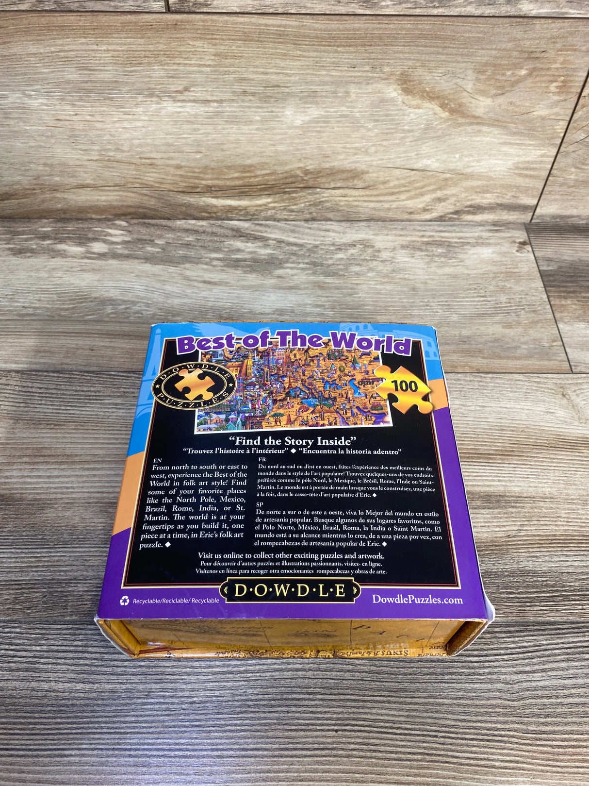 Angled top-down view of Dowdle Best of the World 100-piece jigsaw puzzle box showcasing back artwork and purple border on a wood tabletop