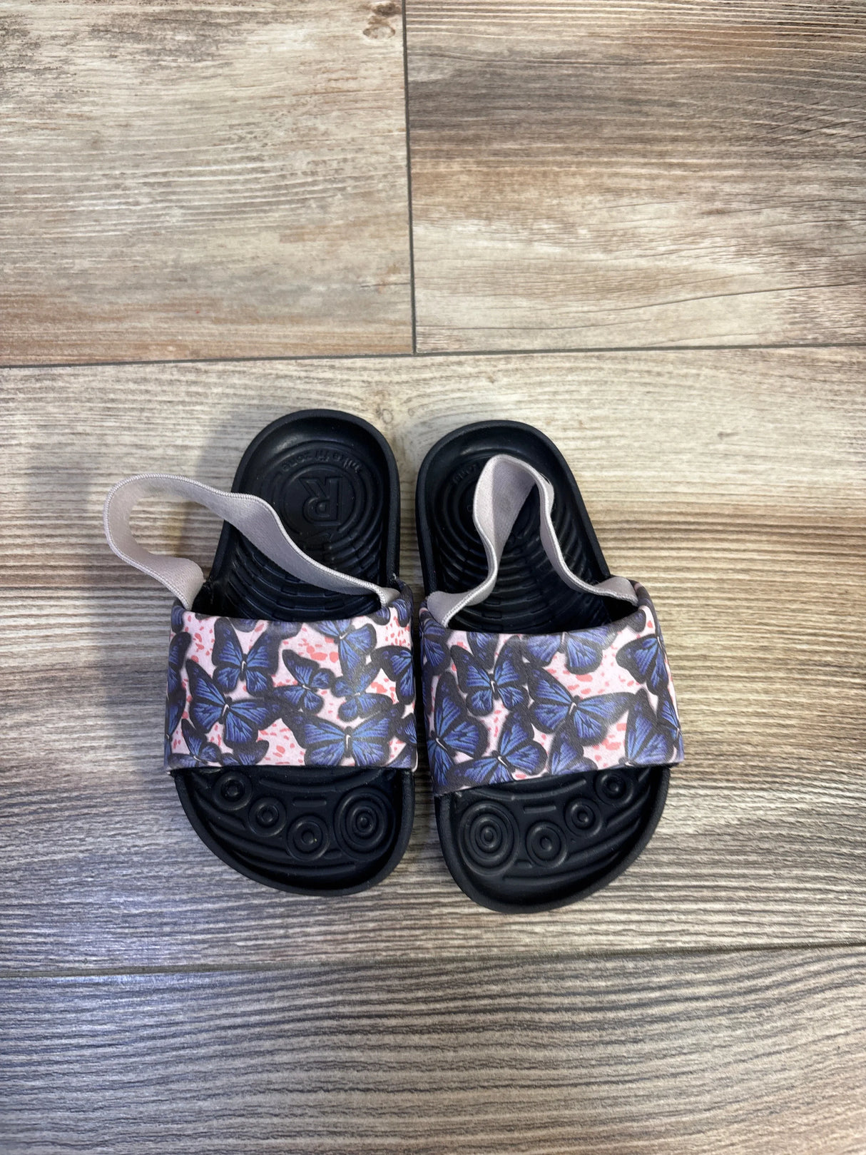 Top-down view of toddler Nike Kawa Slide sandals showcasing pink butterfly-printed strap and black textured footbed on wood floor