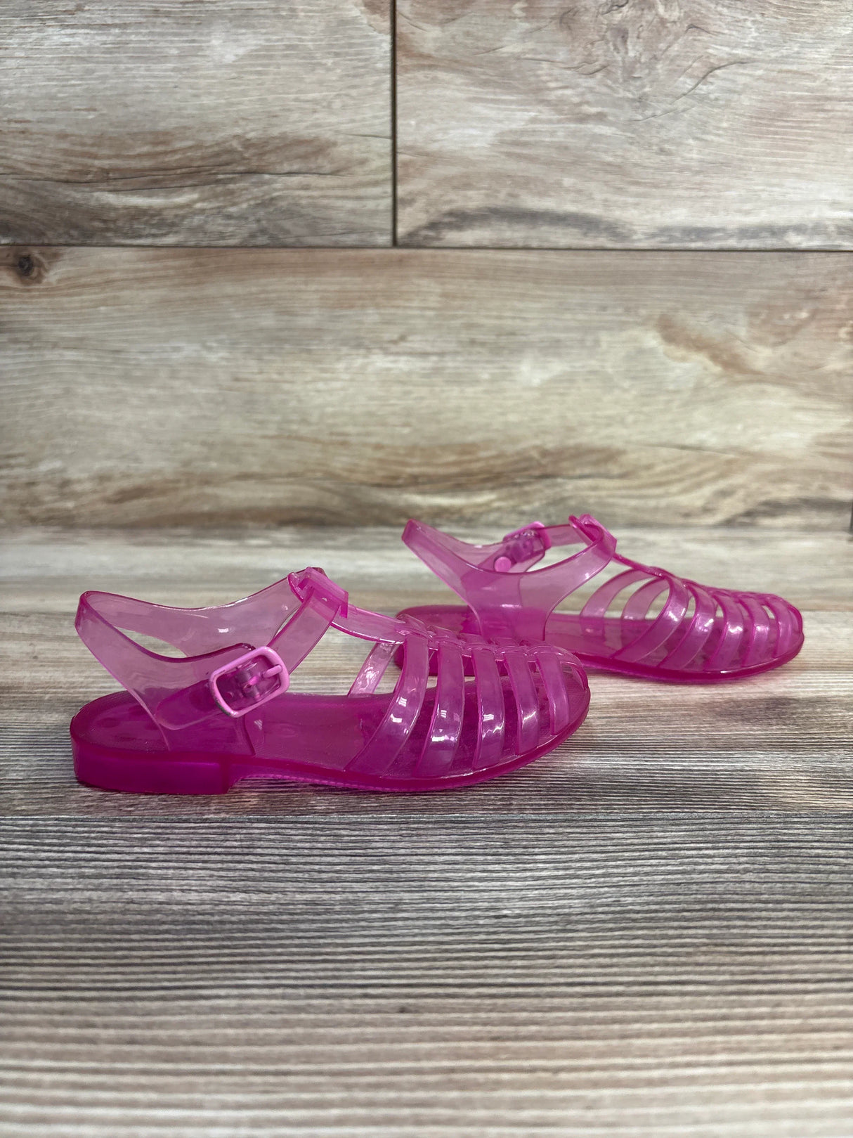 Side view of Janie and Jack Jelly Fisherman Sandals in translucent pink showcasing caged upper and buckle ankle strap on wood plank floor