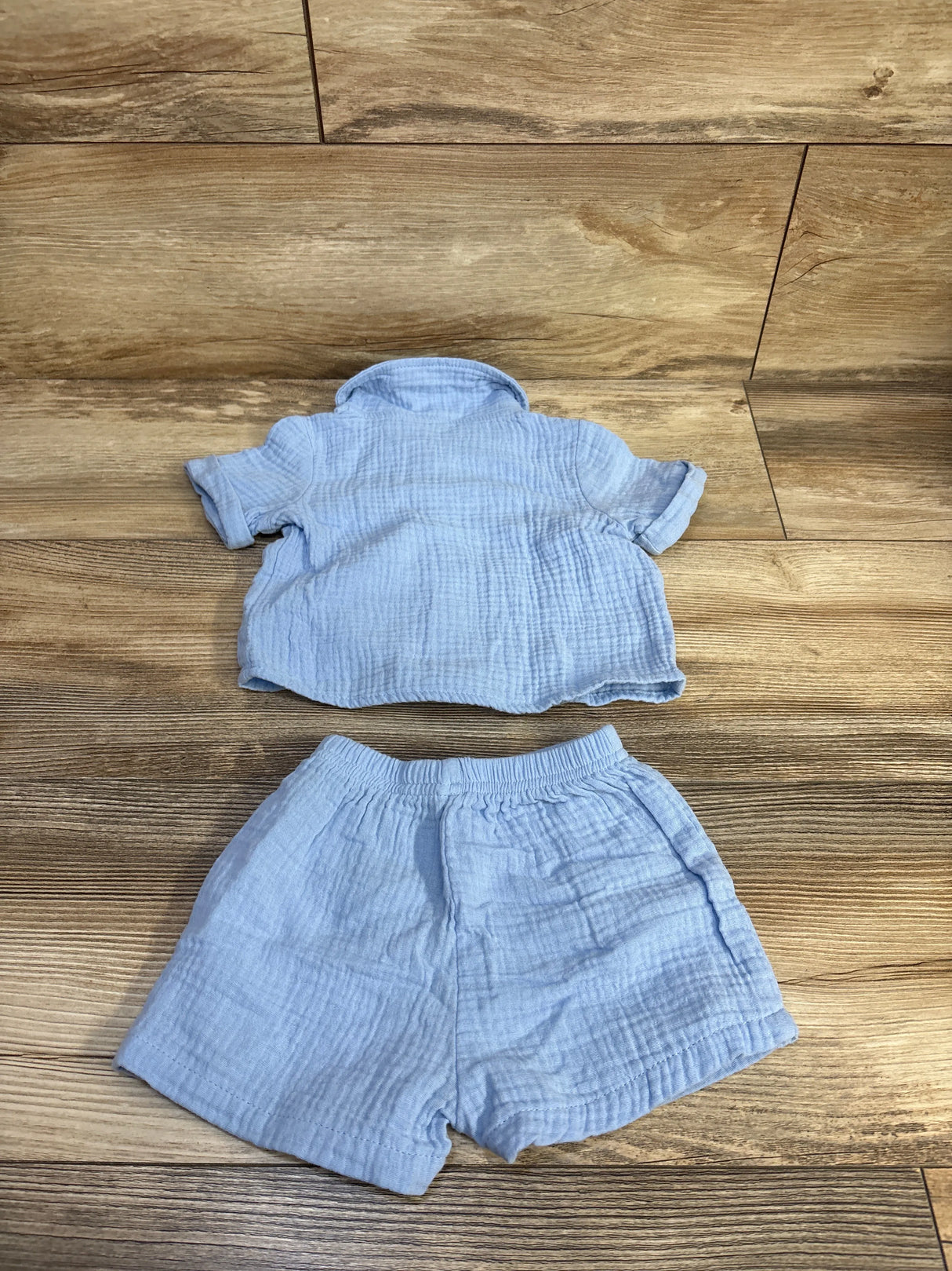 Flat lay of light blue gauze baby button-up shirt and matching elastic-waist shorts showcasing crinkled texture on wooden floor