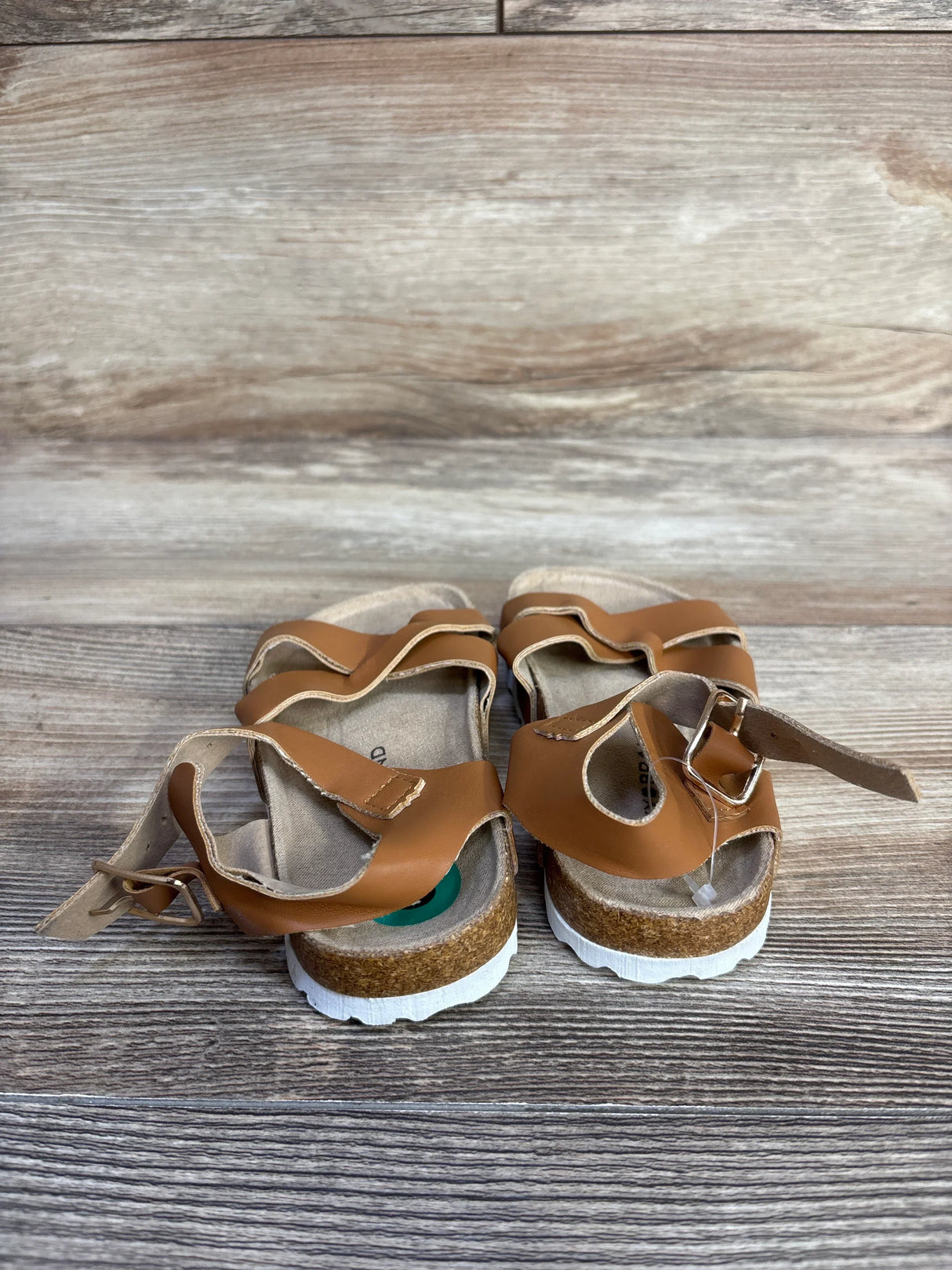Rear view of Lucky Brand Kiara toddler sandals in brown showcasing cork footbed, adjustable buckles, and white ridged soles on wood background