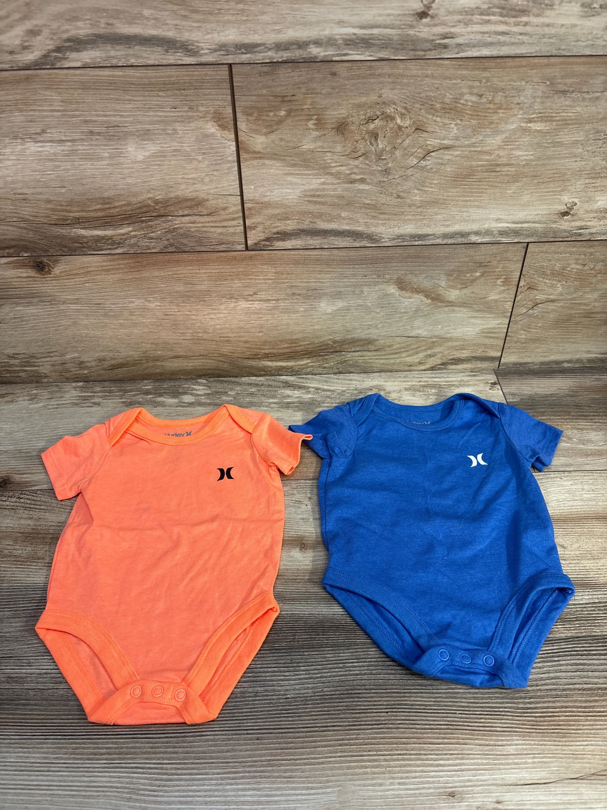 Flat lay of two Hurley 2-pack bodysuits, orange and blue, showing front chest logos and snap closures on wood background