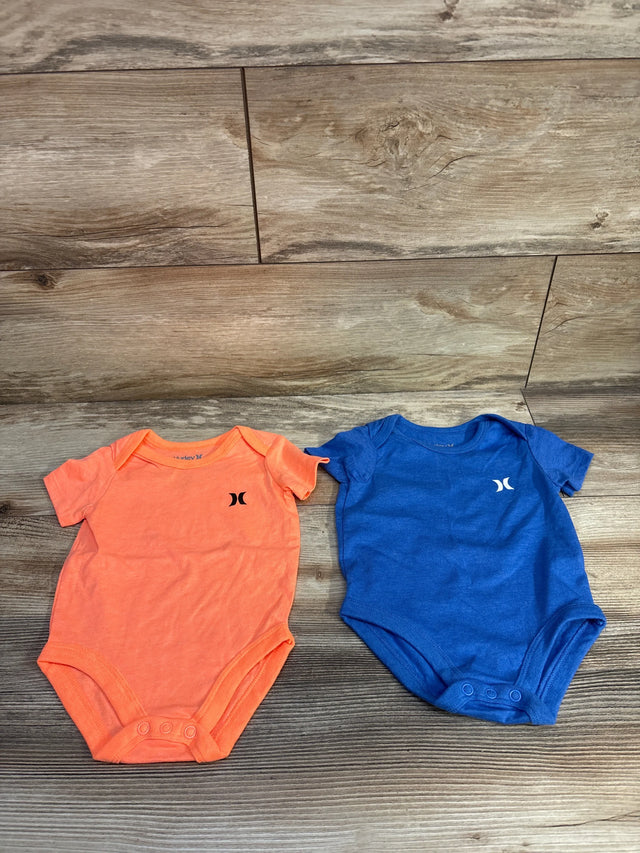 Flat lay of two Hurley 2-pack bodysuits, orange and blue, showing front chest logos and snap closures on wood background