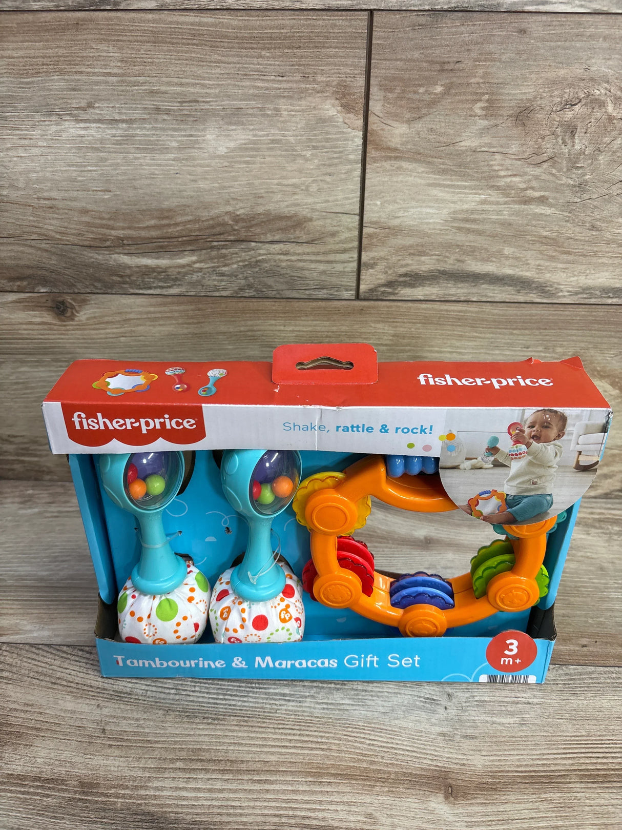 Front view of Fisher-Price tambourine and maracas gift set showing orange tambourine and aqua maracas in box on wood