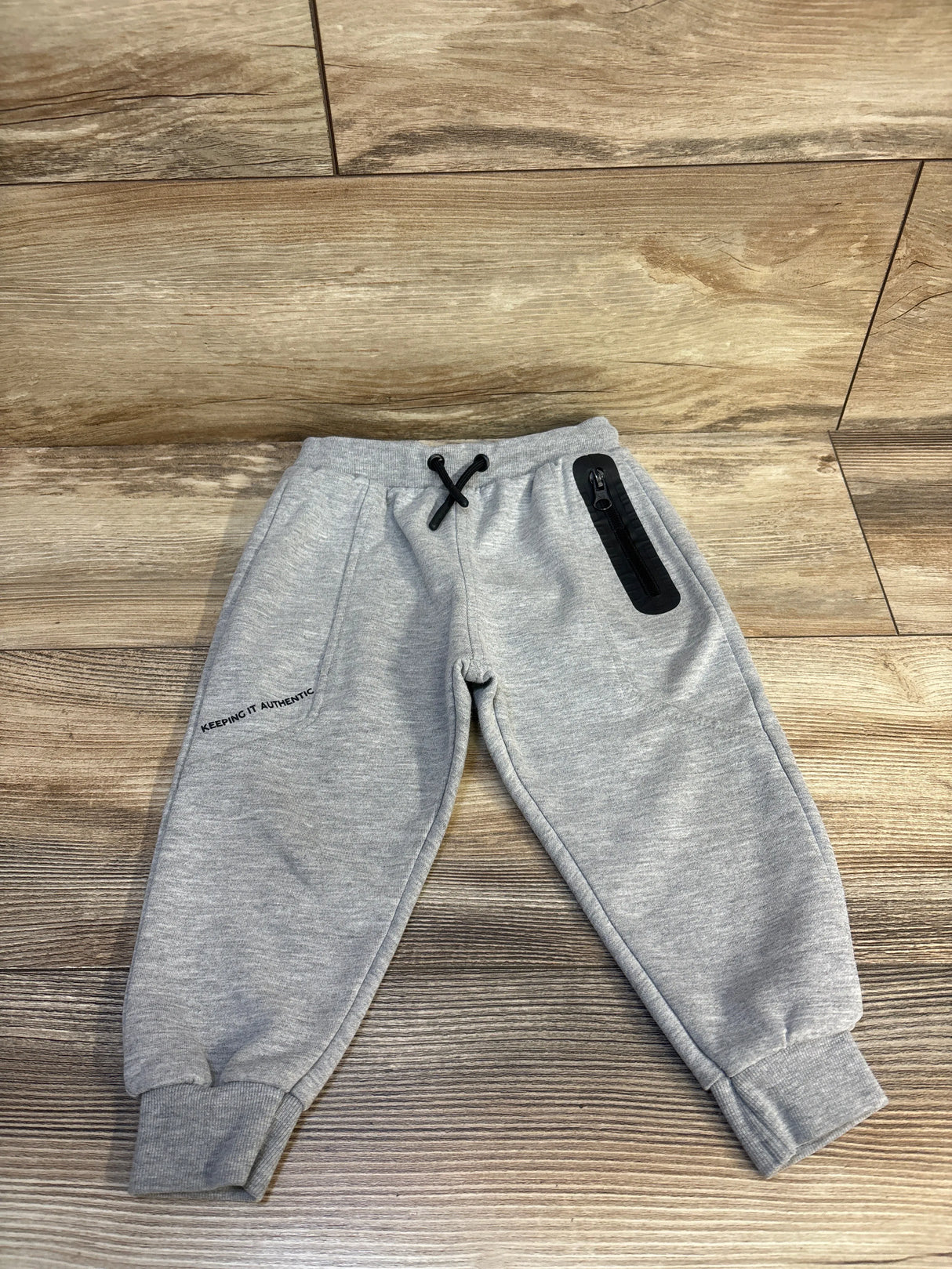 Gray kids joggers with 'Keeping It Authentic' text, black drawstring, and zip pocket on wood floor