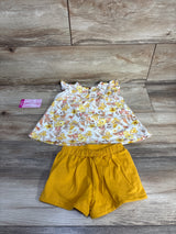 Flat lay of Tommy Bahama baby 2-piece set: white floral flutter-sleeve top and mustard yellow elastic shorts on wood background