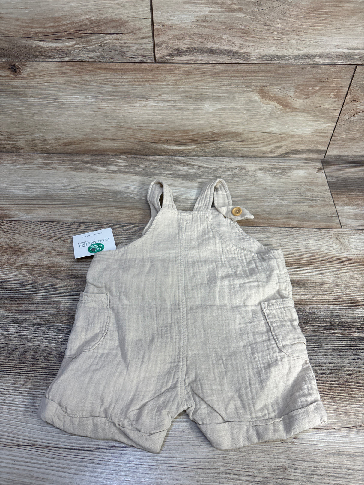 Flat lay of beige Little Planet organic gauze baby shortalls (9m) showing front patch pockets and buttoned shoulder straps on wood floor background