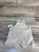 Flat lay of beige Little Planet organic gauze baby shortalls (9m) showing front patch pockets and buttoned shoulder straps on wood floor background