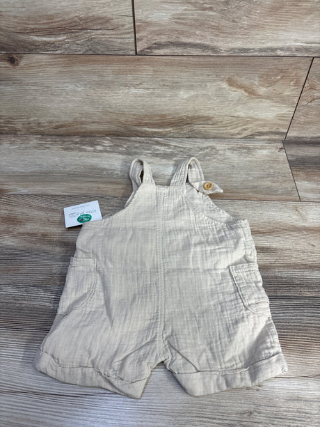 Flat lay of beige Little Planet organic gauze baby shortalls (9m) showing front patch pockets and buttoned shoulder straps on wood floor background