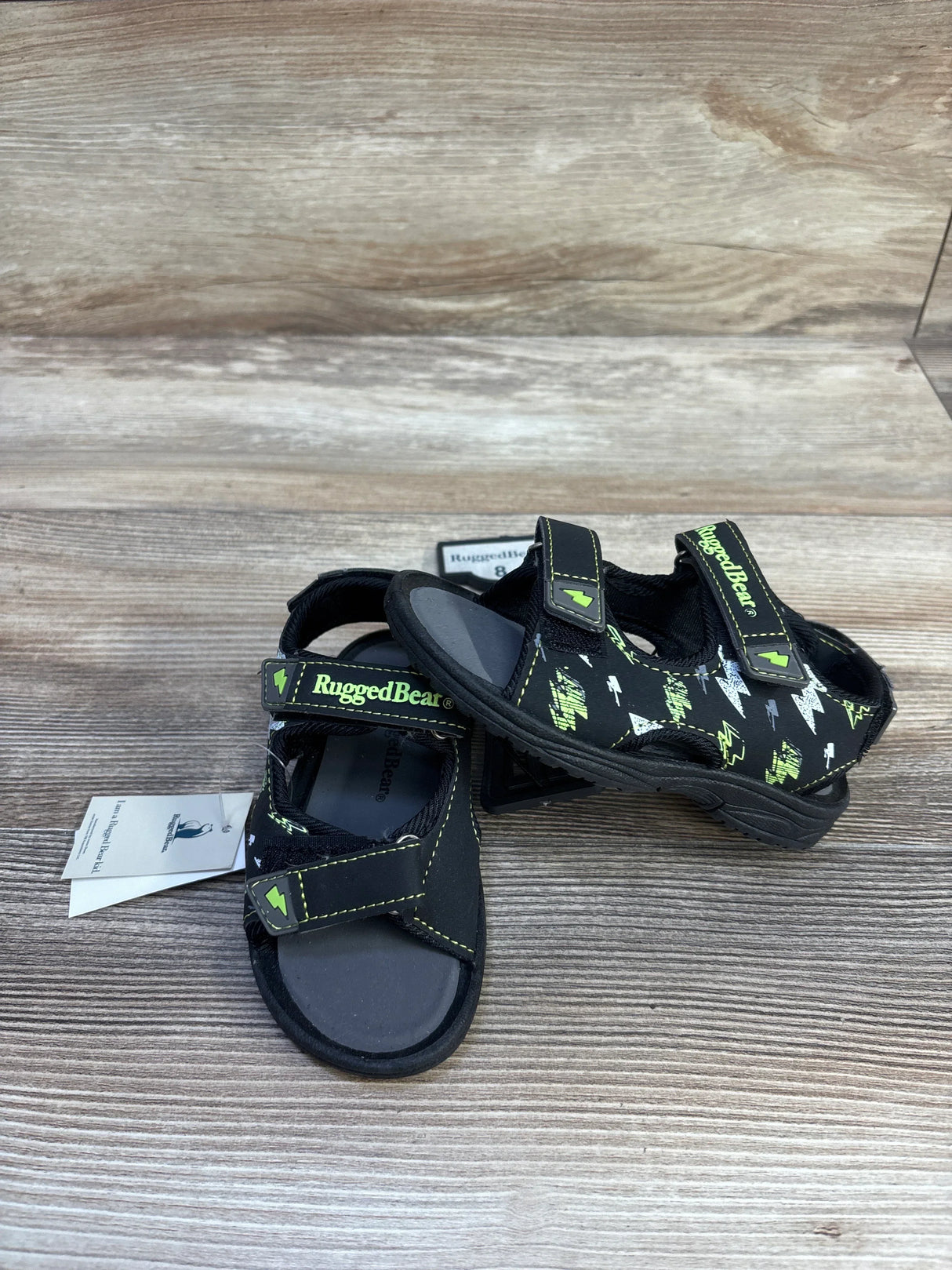 Front three-quarter of Rugged Bear kids' black sandals with green lightning print and Velcro straps on wood background