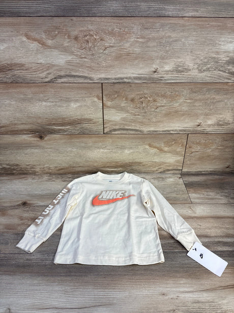 Flat-lay of Nike toddler long-sleeve shirt size 2T showcasing coconut milk cream color, orange swoosh and "Just Do It" sleeve text on wood background