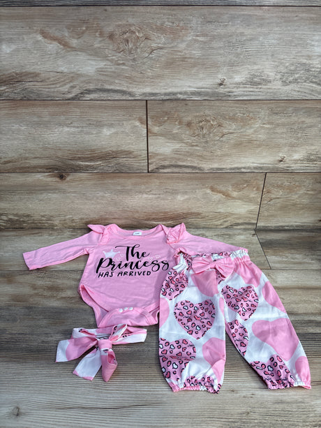 Flat lay of pink 3-piece baby set: bodysuit reading 'The Princess Has Arrived', heart-print pants and matching headband on wood background