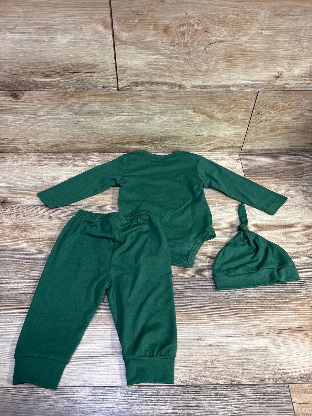 Top-down view of green 3-piece baby bodysuit, pants, and knotted hat set with long sleeves on a wood-look background