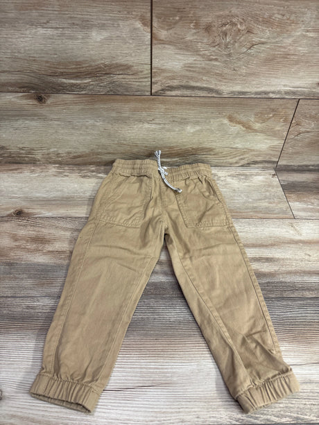 Front view of H&M tan pull-on drawstring joggers, size 3T, with pockets and elastic ankle cuffs on a wood-look background